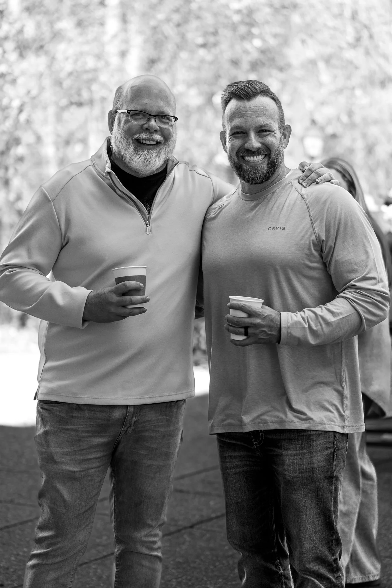 Two men smiling and holding paper cups, standing outdoors with a blurred natural background, one with glasses and a beard, the other with a beard and short hair, with an arm around each other's shoulders.