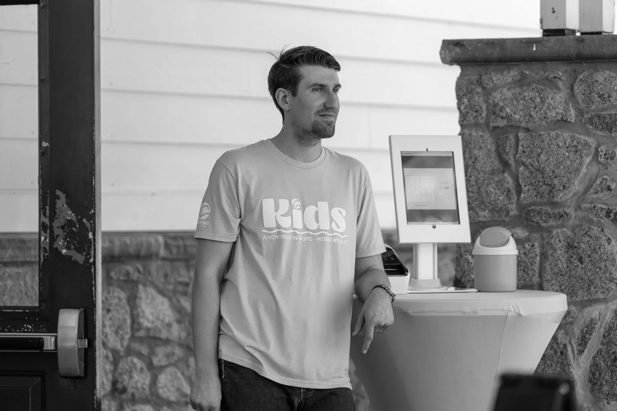 A man in a casual t-shirt standing indoors near a counter with a touchscreen kiosk and a small container on it. Stone wall in background.