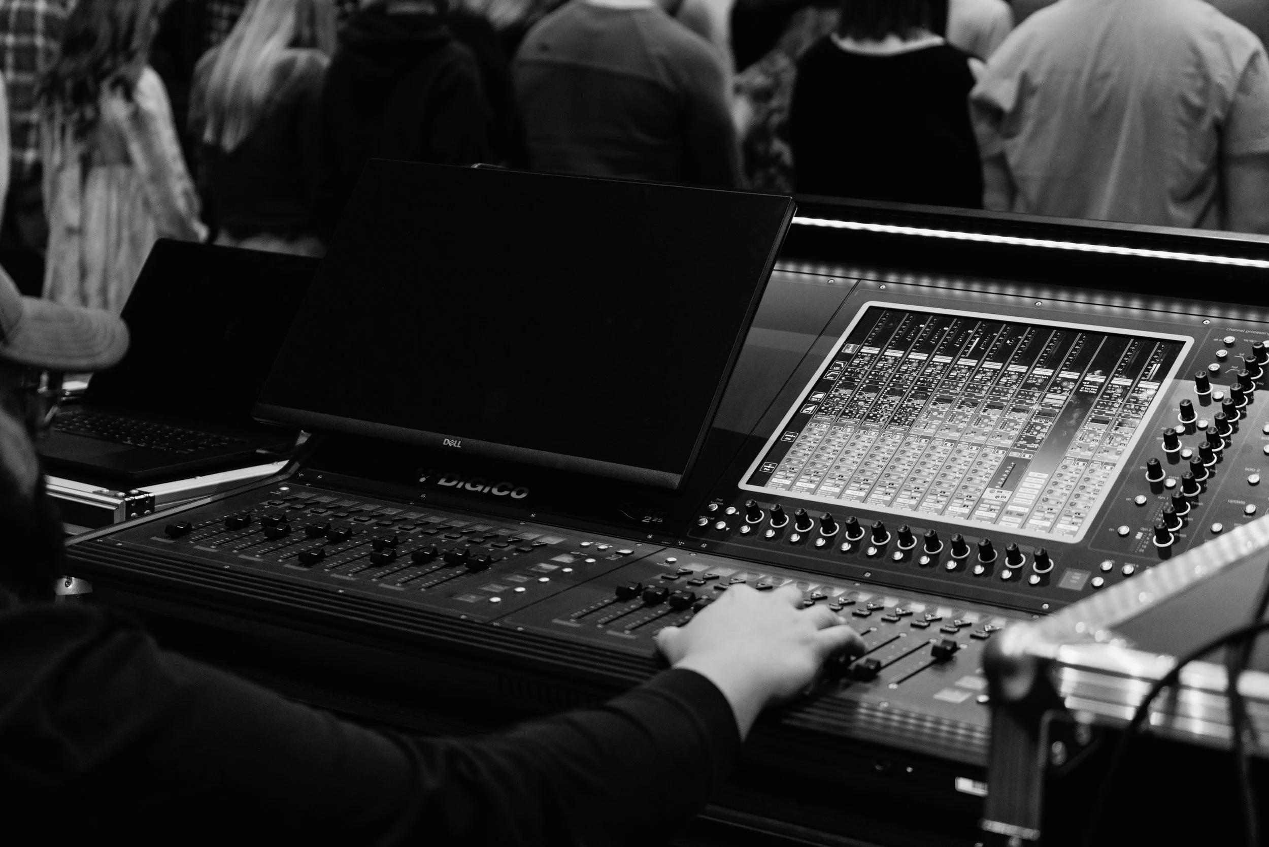 Close-up of a digital sound mixing console with a person's hand adjusting controls, surrounded by people in the background.