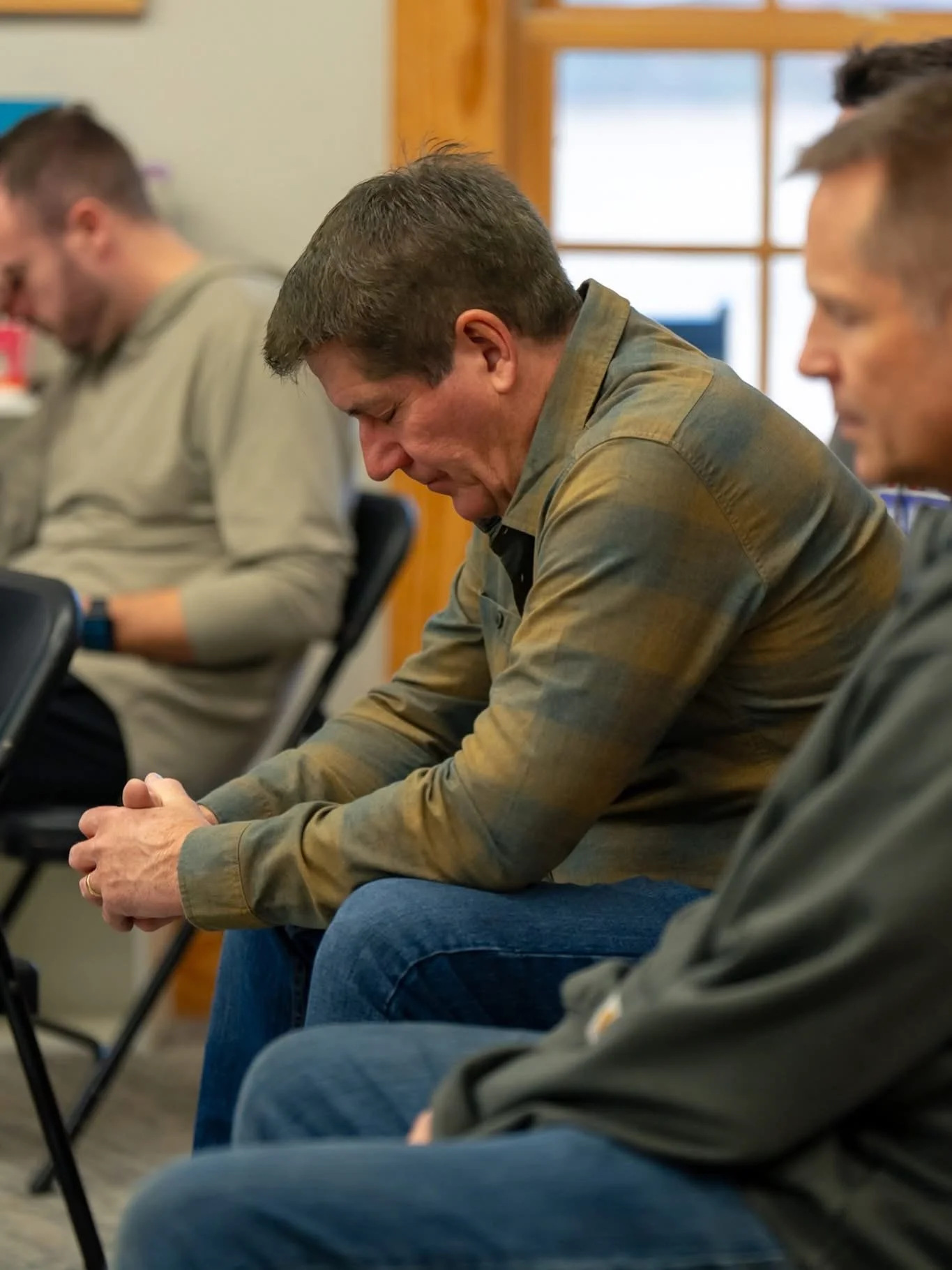 A group of men seated in a room, with one man in the center looking down, hands clasped, appearing to be in prayer or reflection.