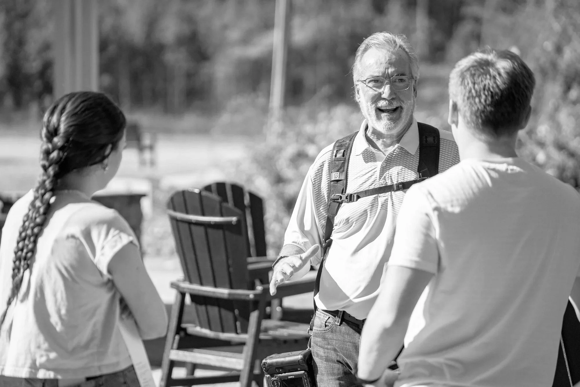 Three people having a conversation outdoors, including an older man with glasses and a backpack, a woman with a long braided hair, and a man with short hair, with chairs stacked behind them.