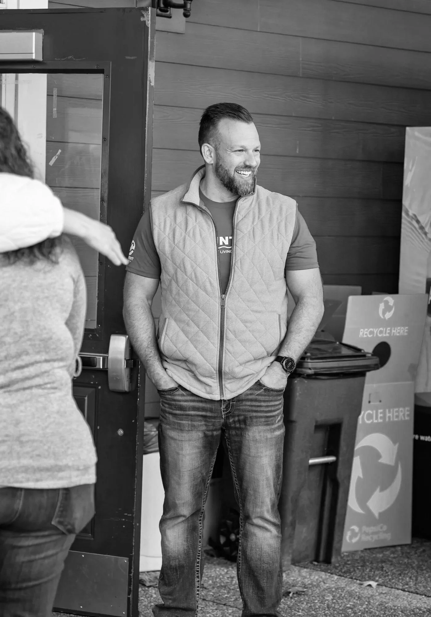 A man with a beard and short hair smiling, standing with his hands in his pockets in front of a wall with wood paneling, wearing a zip-up vest over a t-shirt.