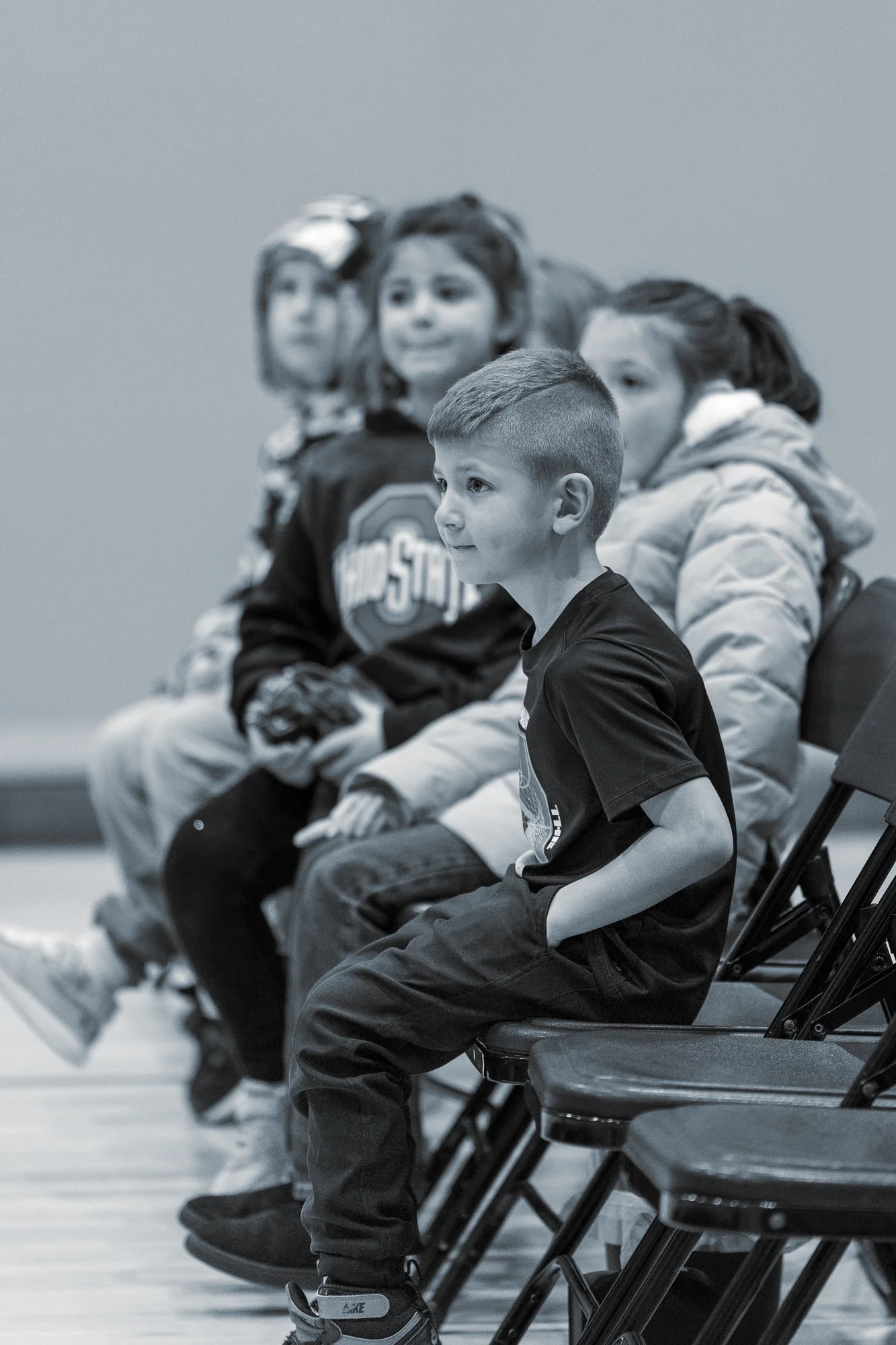 Young boy sitting on a folding chair, watching attentively, with other children sitting behind him.