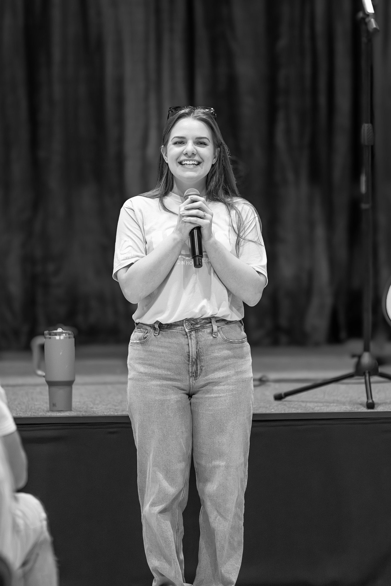 A woman standing on stage holding a microphone, smiling, with a dark background and a tumbler on a table nearby.