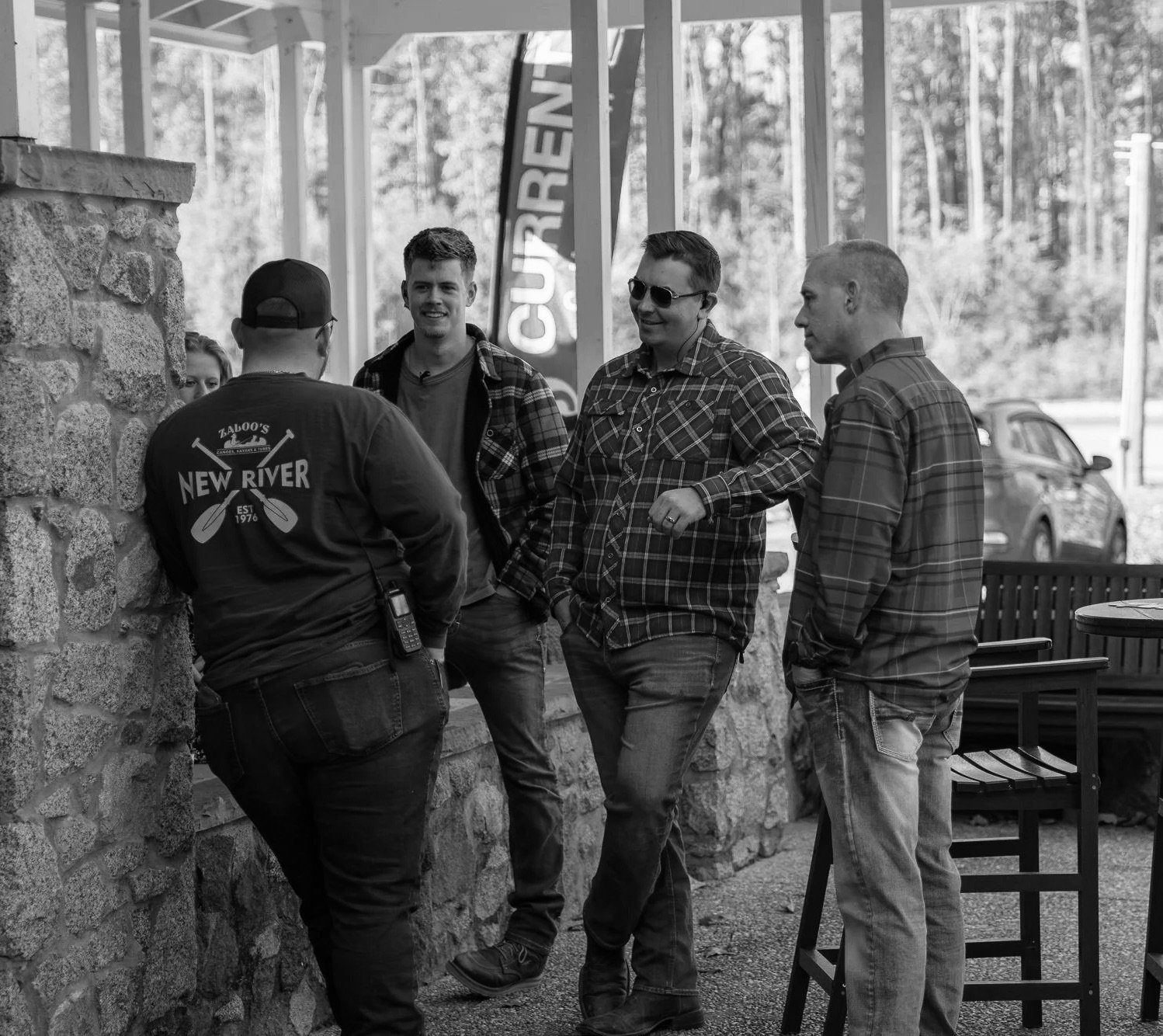 Group of four men casually talking outdoors near a stone wall and a wooden bench, with a car and trees in the background.
