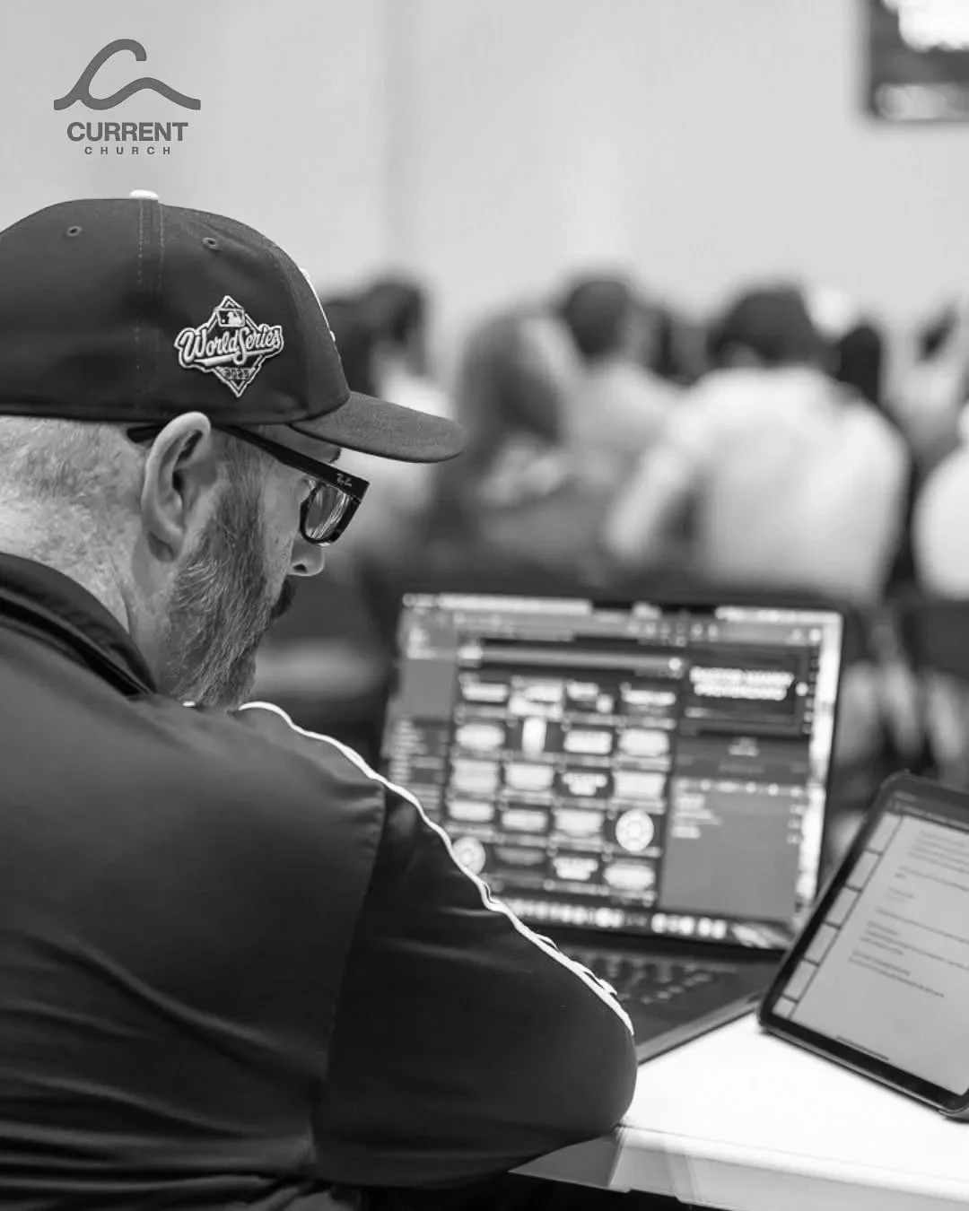 A man wearing glasses, a cap with a logo, and a dark jacket is working on his laptop with multiple screens, in a room with blurred people in the background.