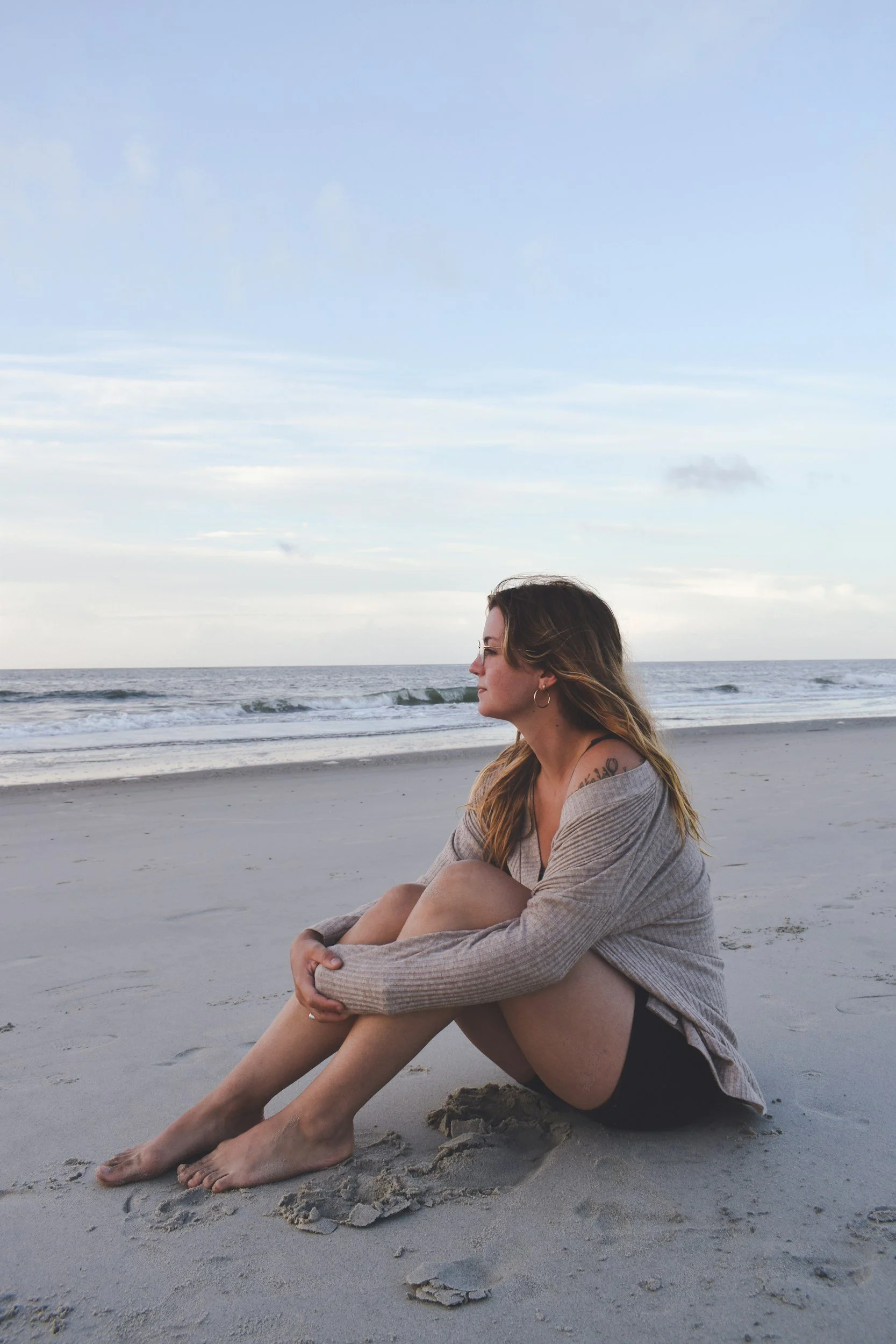 A woman sitting on the beach with her knees pulled up, gazing at the ocean, wearing a beige sweater and black shorts, with a tattoo visible on her shoulder, under a cloudy sky.