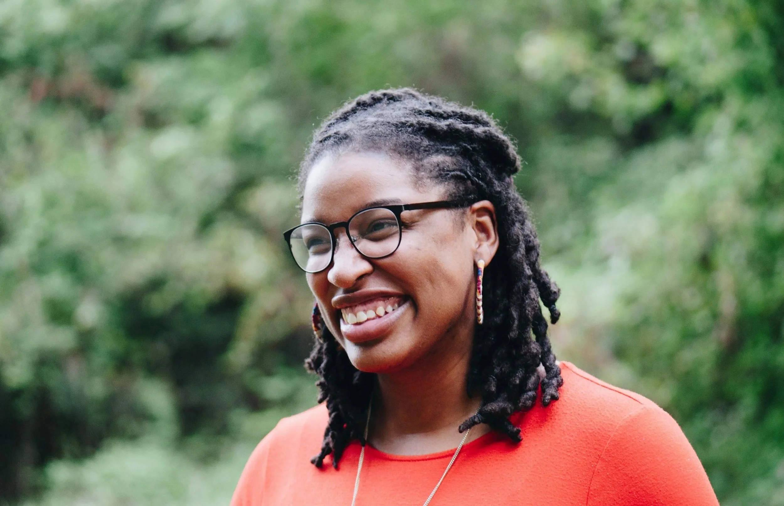 A woman with dreadlocks, wearing glasses, earrings, and an orange top, smiling outdoors with a green, blurry background.