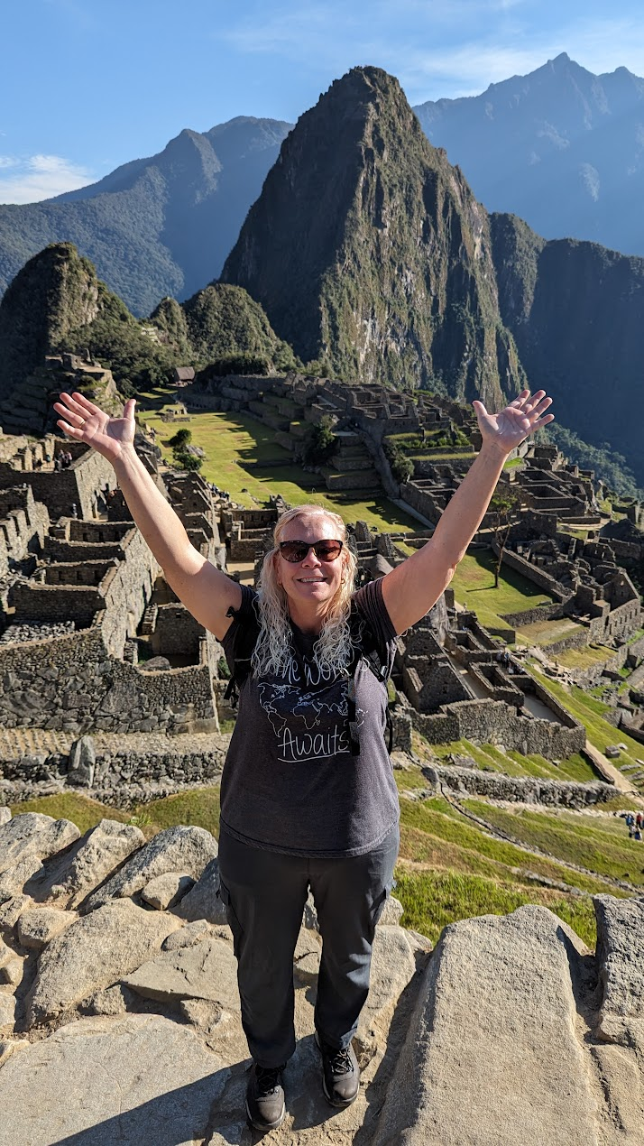 Woman with sunglasses and gray hair celebrating with arms raised at Machu Picchu, ancient Incan ruins, with steep mountain and lush landscape in the background.
