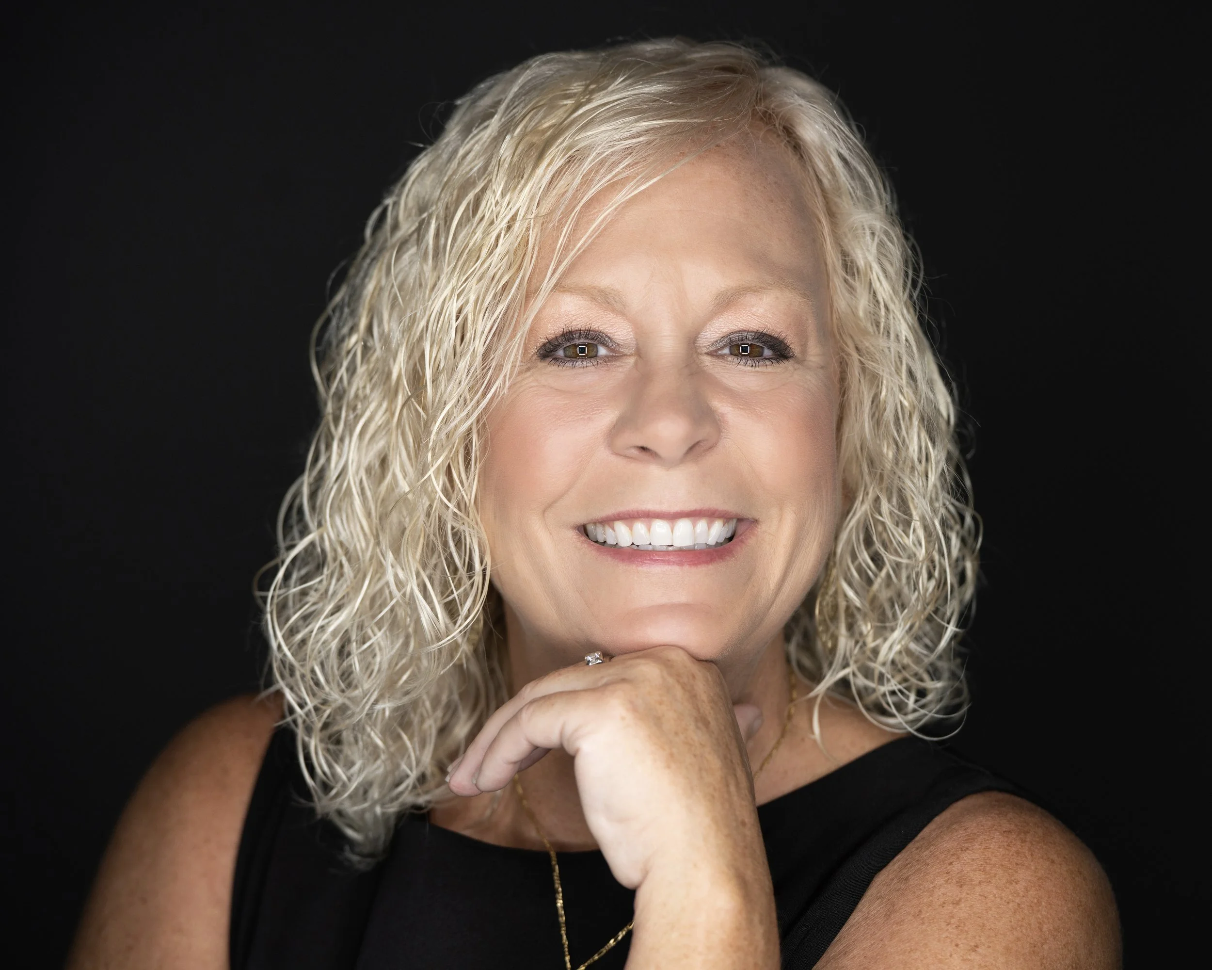 A smiling woman with curly blonde hair, wearing a black top, resting her chin on her hand, against a black background.