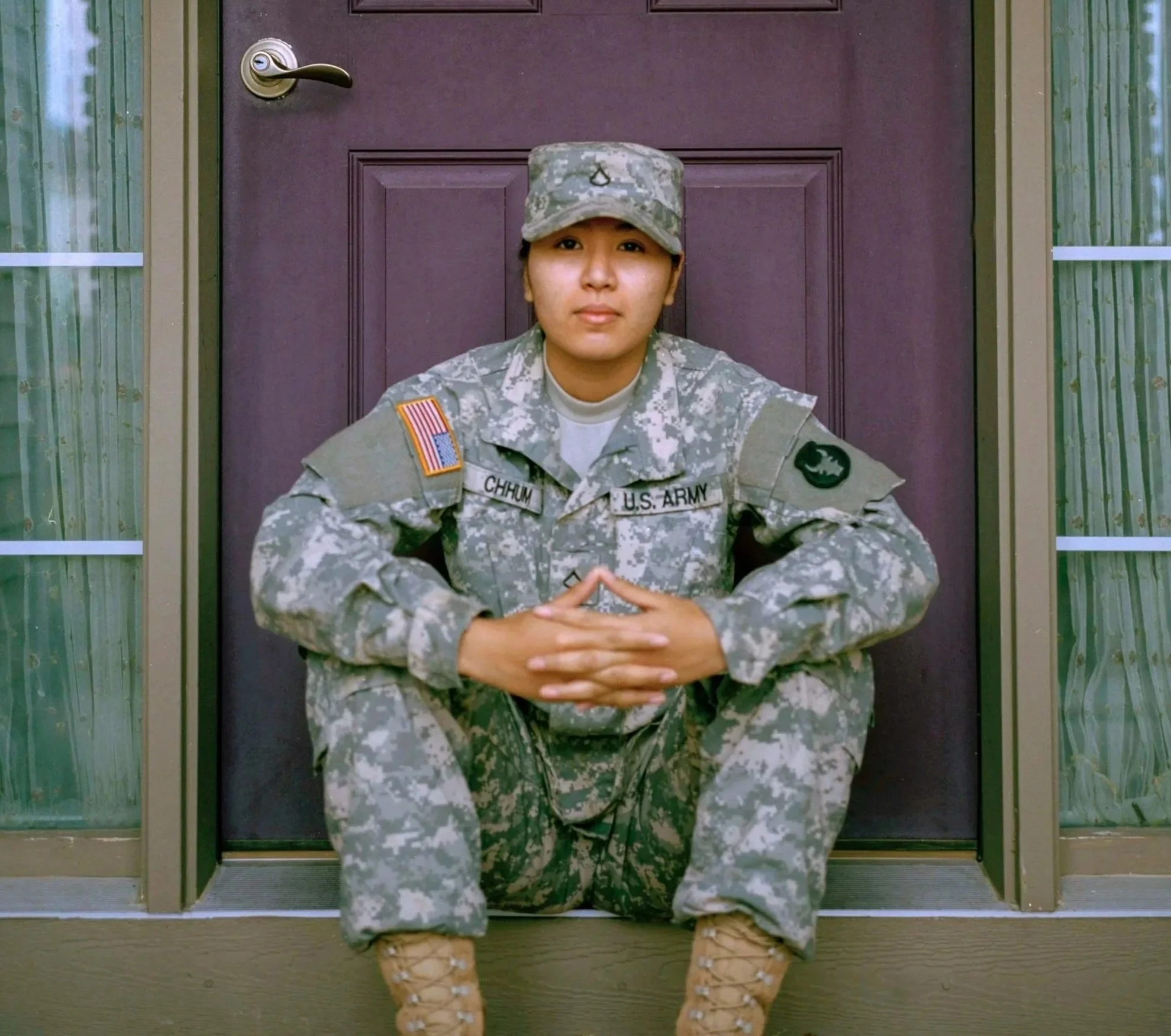 A young woman in military uniform sitting in front of a purple door with hands clasped on her knees.