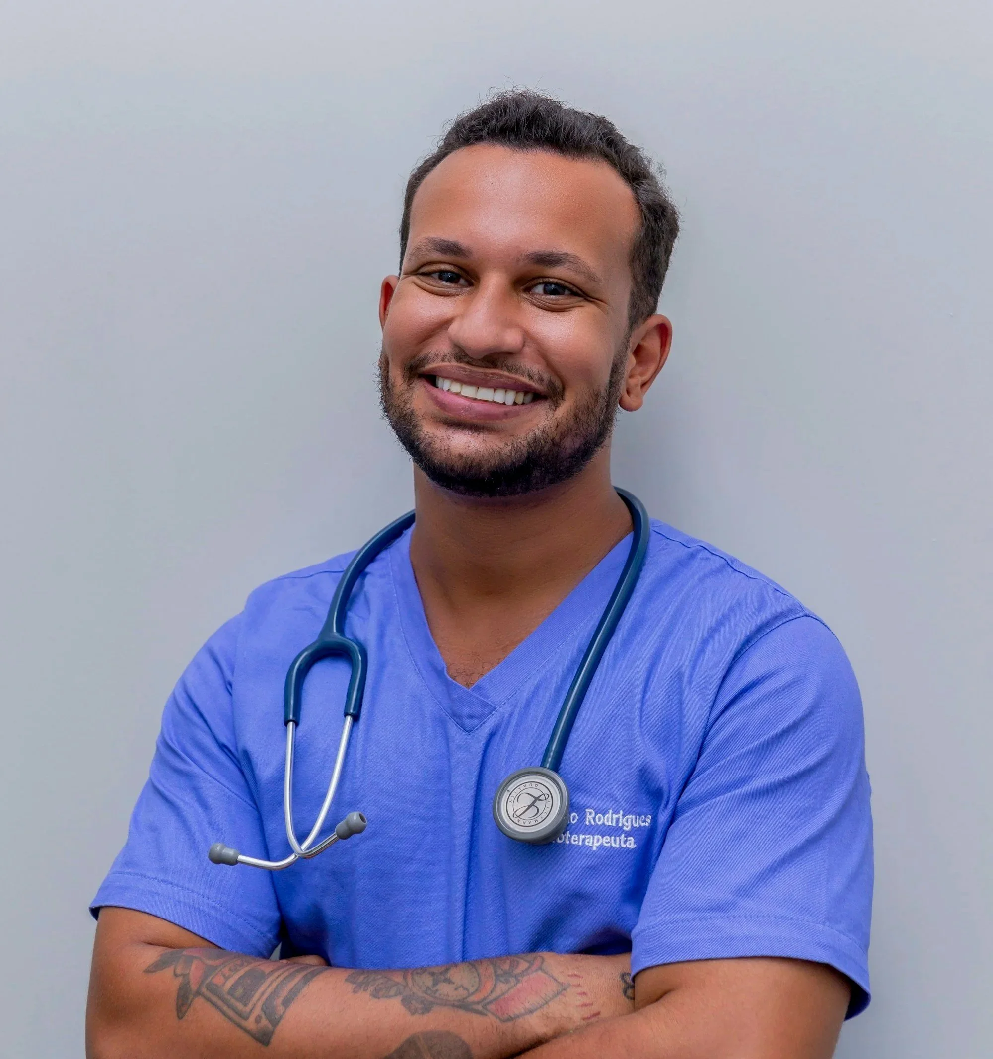 A smiling male healthcare professional wearing blue scrubs and a stethoscope around his neck, standing against a plain wall.