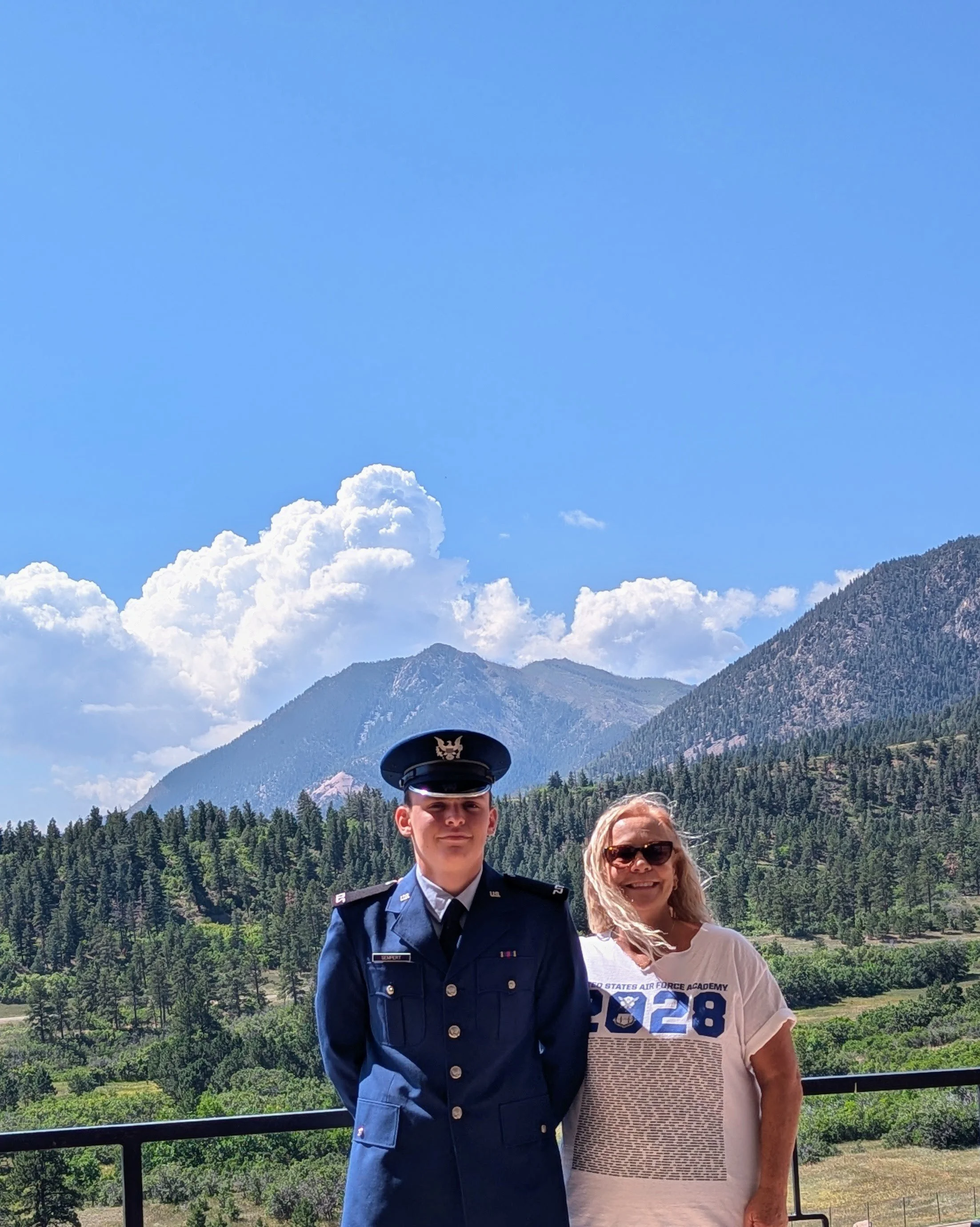 A young man in a military uniform and a woman in sunglasses standing outdoors with mountains and a blue sky in the background.