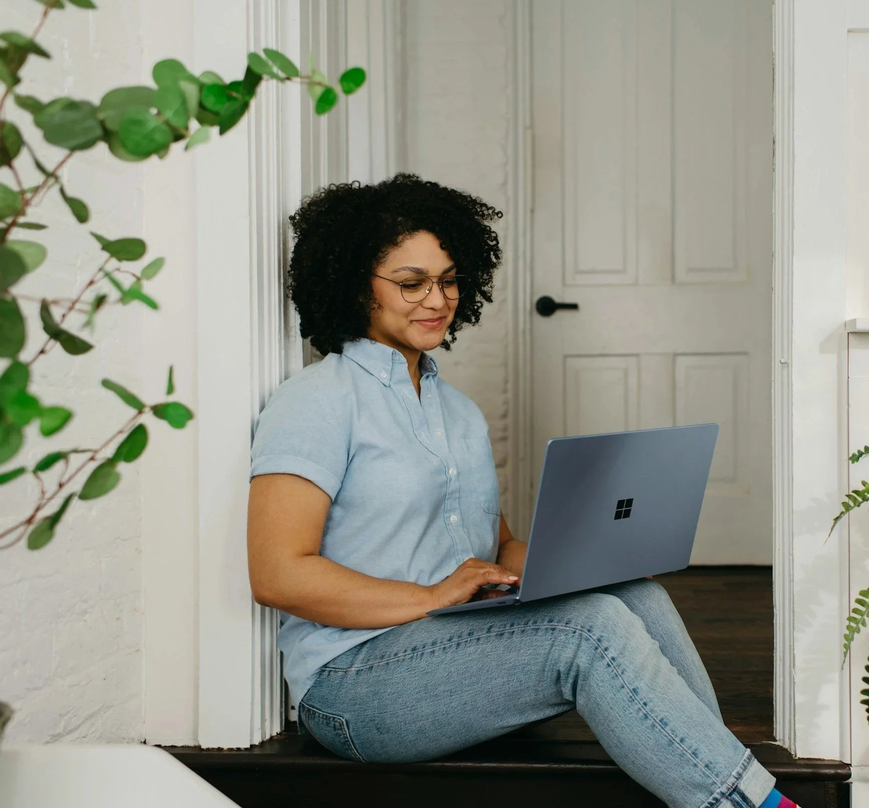Woman with curly hair and glasses sitting on the floor near a doorway, working on a gray Microsoft Surface laptop.
