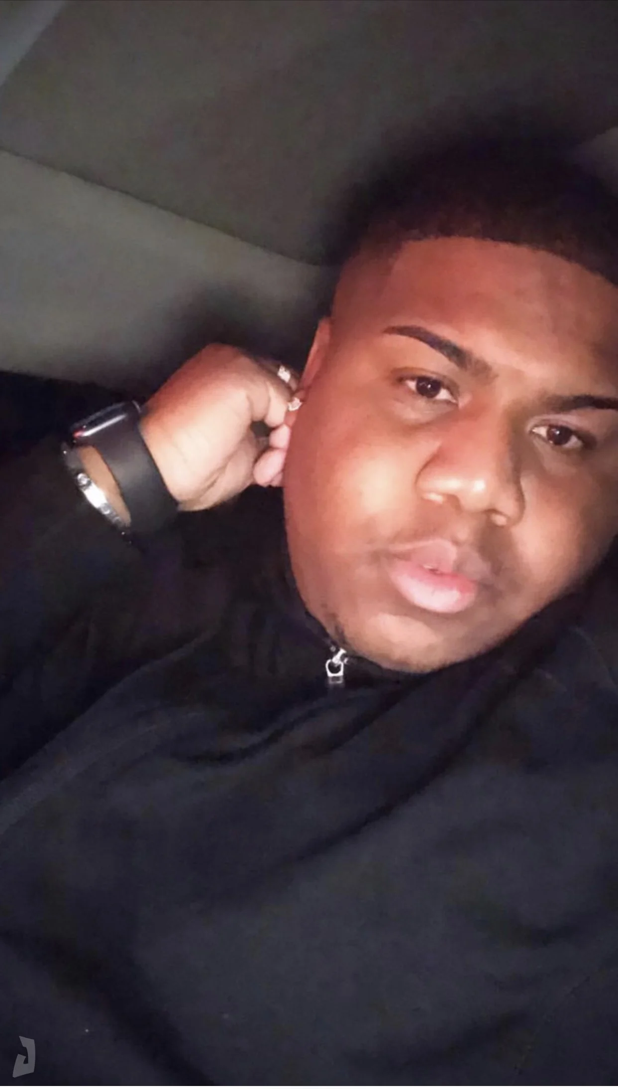 A young man with short dark hair, wearing a black hoodie, lying down with his hand touching his ear, looking at the camera.