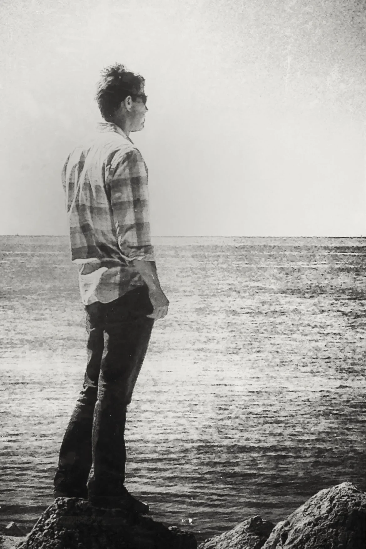 A black and white photograph of a person standing on rocks by the water, facing the ocean, wearing a checkered shirt and sunglasses.