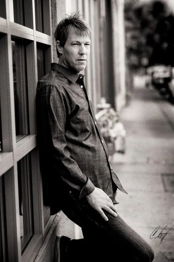 A man leaning against a wall outdoors in black and white, wearing a button-up shirt and jeans, looking at the camera.