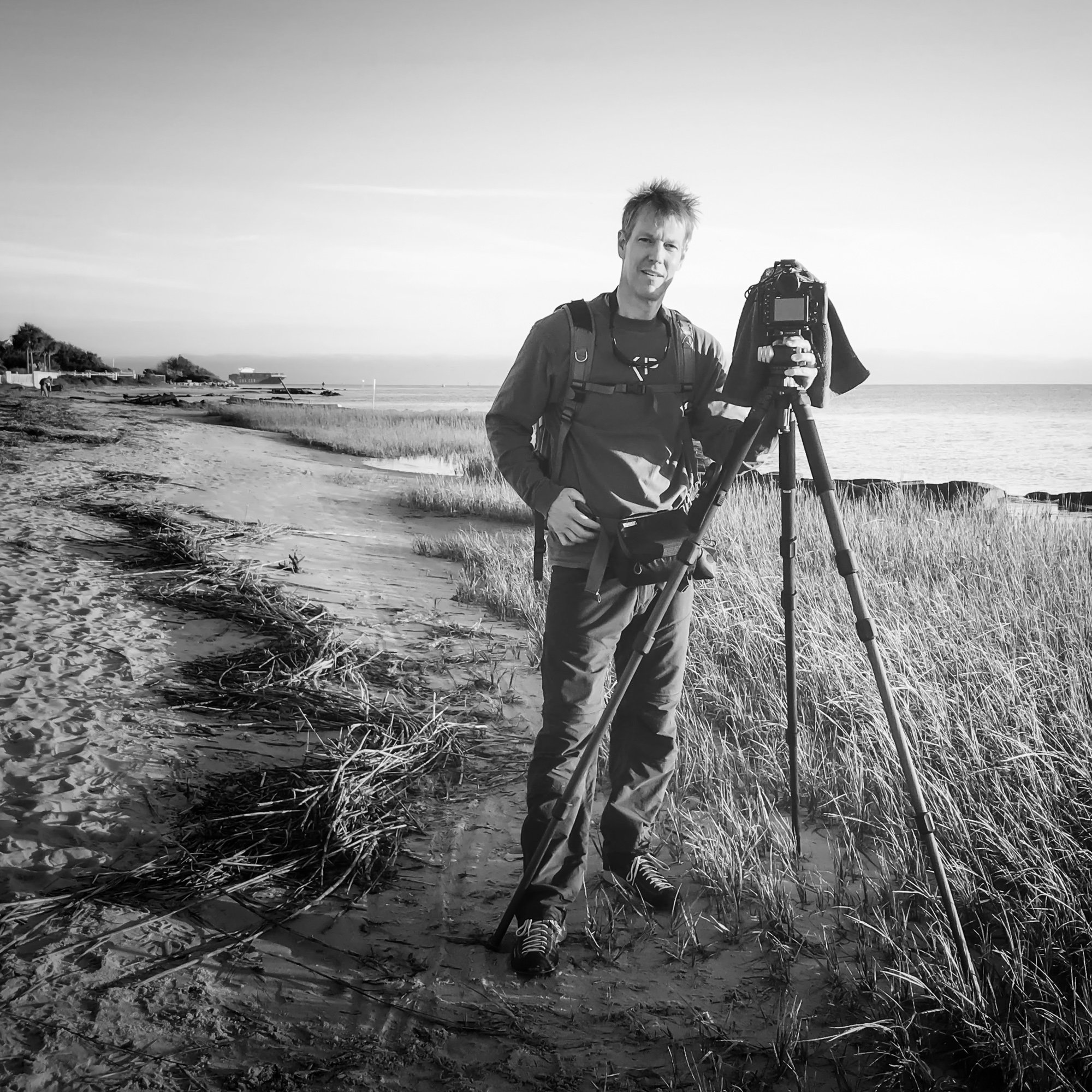 A man with a backpack standing on a beach holding a camera on a tripod.