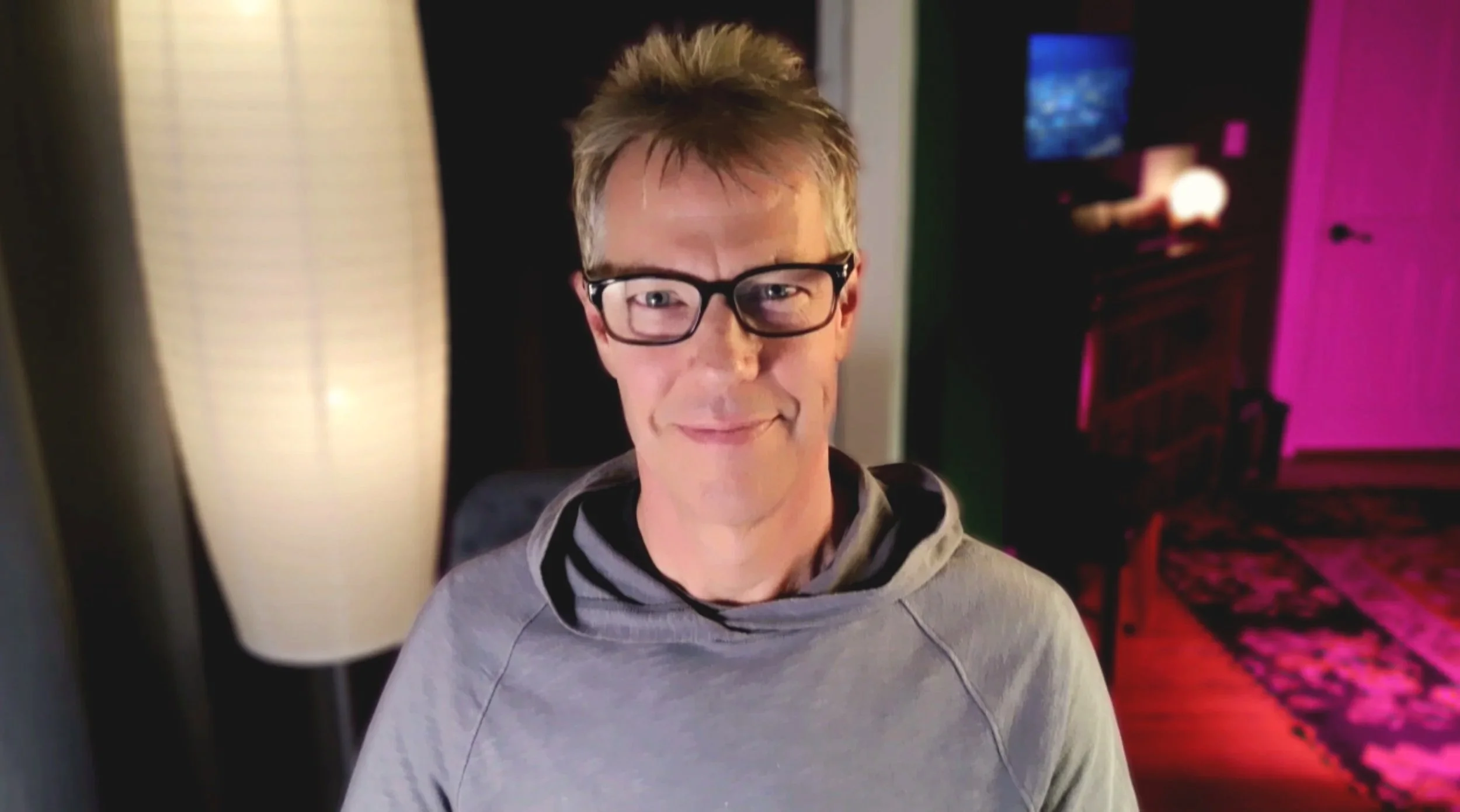 A man with glasses wearing a gray hoodie smiling in a room with a lamp and a colorful background.