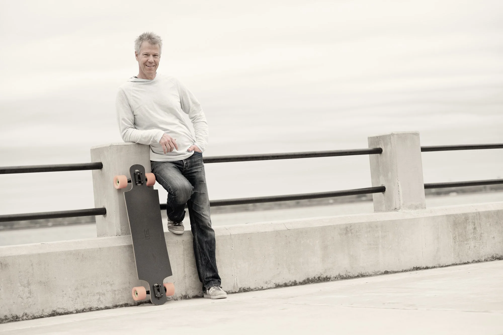 Kevin Holliday wearing a light blue hoodie and jeans leaning against a concrete barrier at the Charleston Battery, with a skateboard propped against the barrier beside him, on an overcast day.