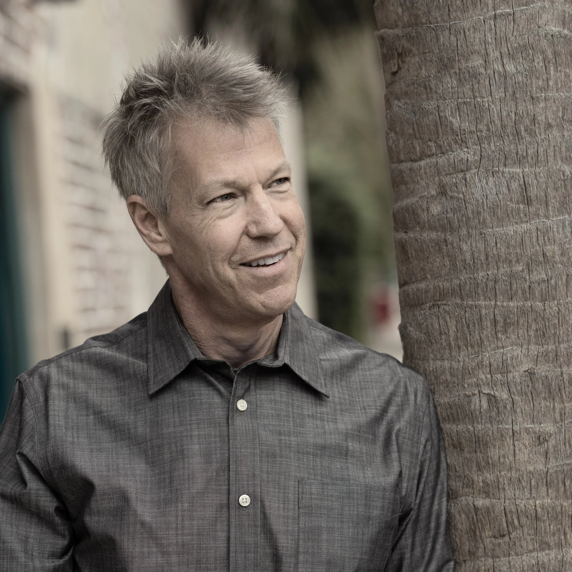 Close-up of Kevin wearing a gray shirt, standing outdoors next to a palm tree.