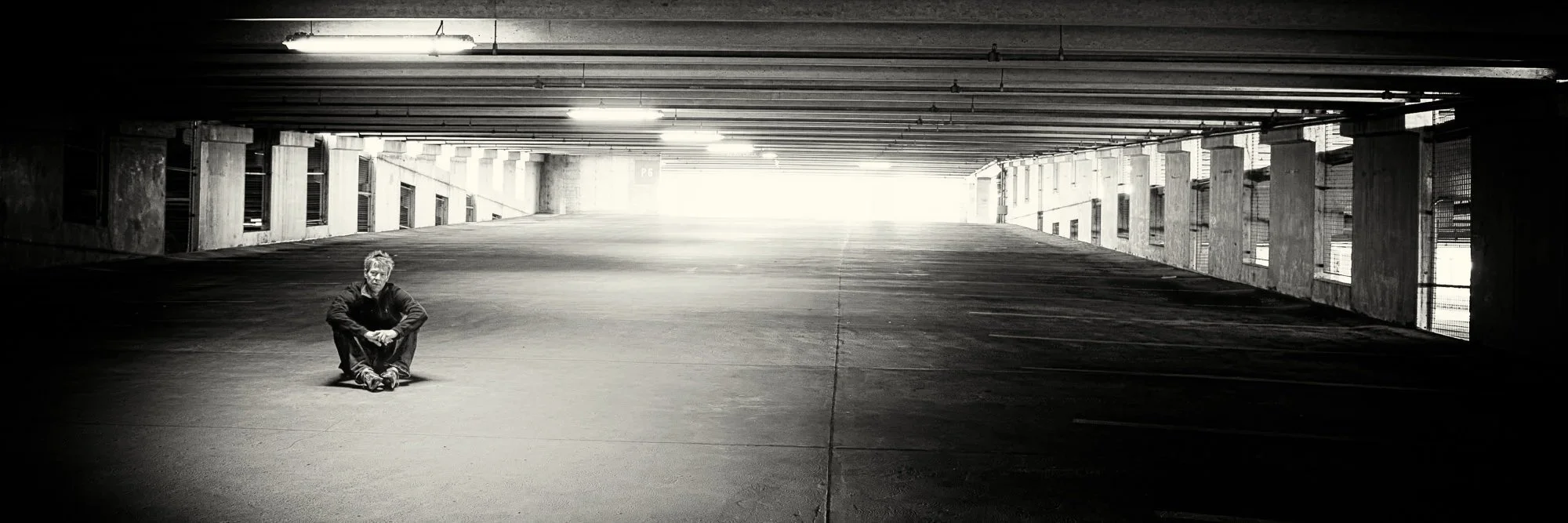 A man sitting on the ground in an empty, dimly lit parking garage with bright light at the far end of the space.