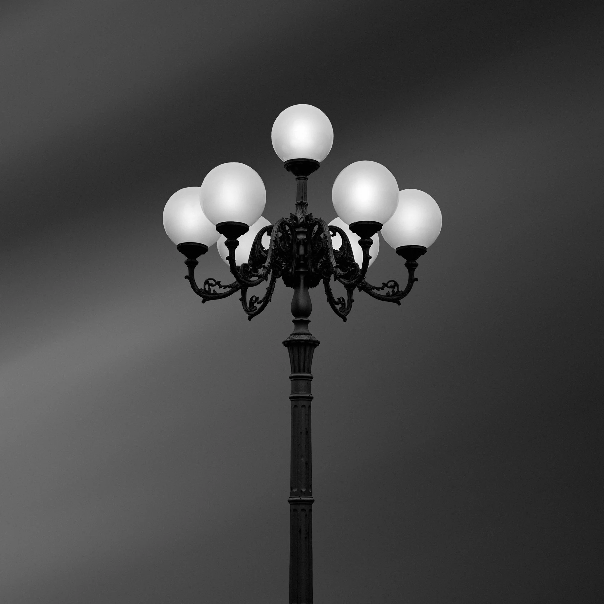Black and white photo of a vintage street lamp with eight spherical lights on ornate curved arms, against a cloudy sky.
