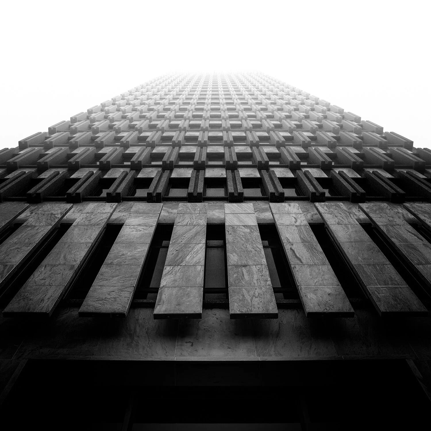 Black and white photo of a tall modern skyscraper viewed from the ground, stretching into the foggy sky.