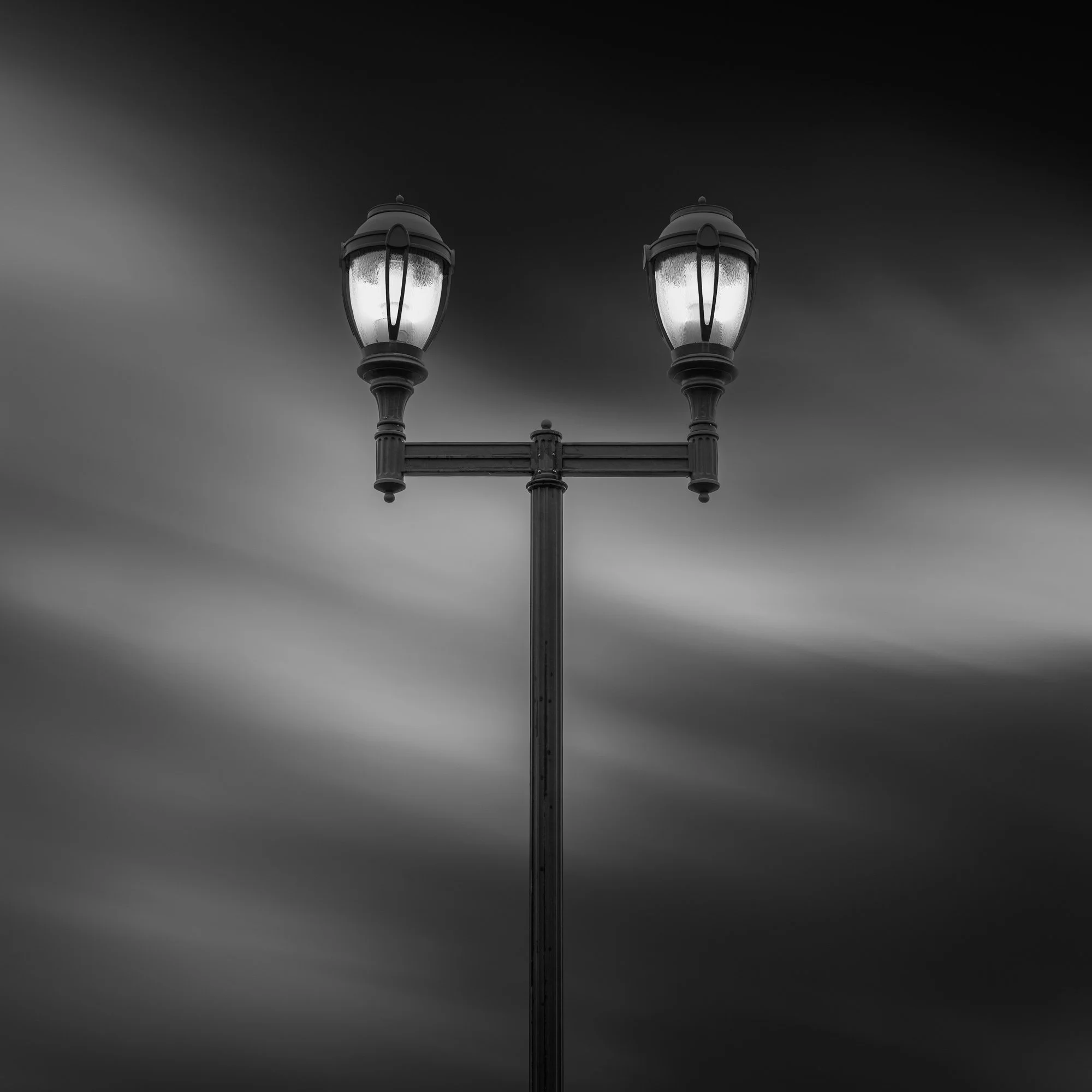 Black and white photo of a vintage street lamp with two lit lanterns against a cloudy sky.