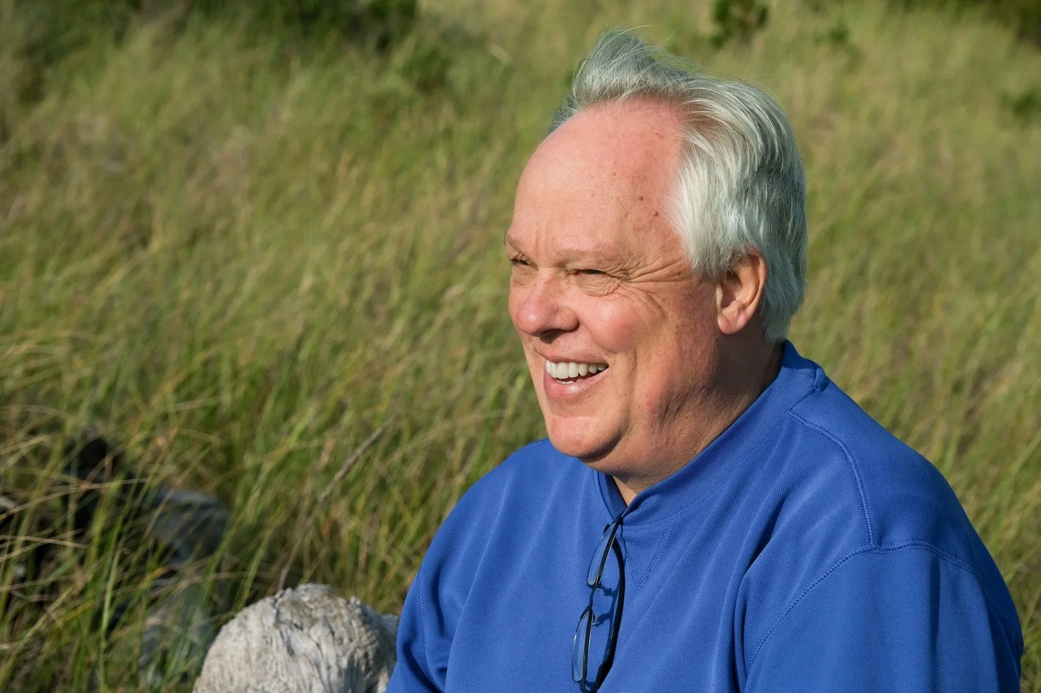 A friendly, approachable man smiling outdoors, sitting on a grassy field.