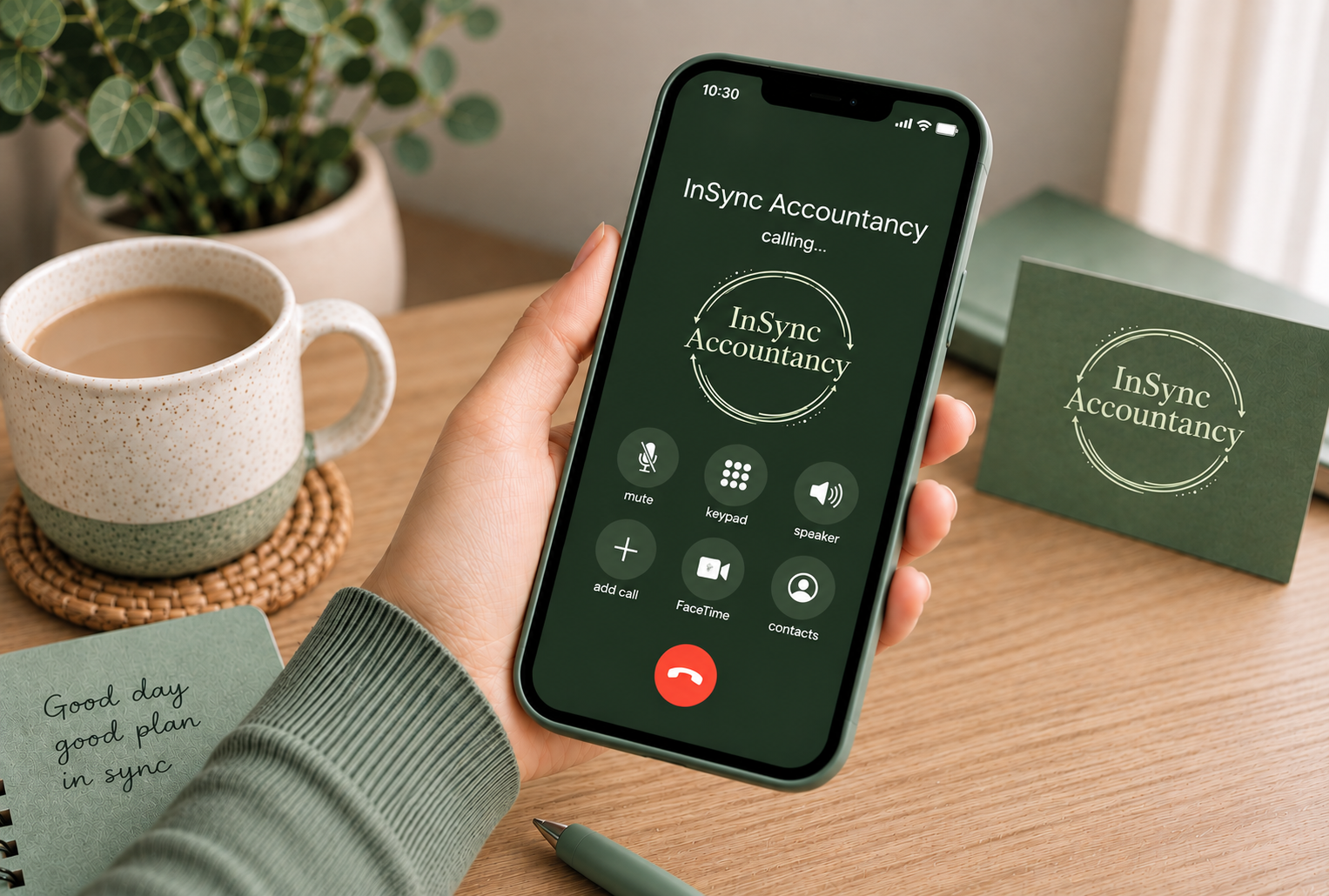 A person holding a smartphone during a video call with an app called 'InSync Accountancy.' The phone is on a wooden desk with a coffee mug, a small notebook with the message 'Good day good plan in sync,' a pen, and a business card with the same 'InSync Accountancy' logo. In the background, there is a potted plant.