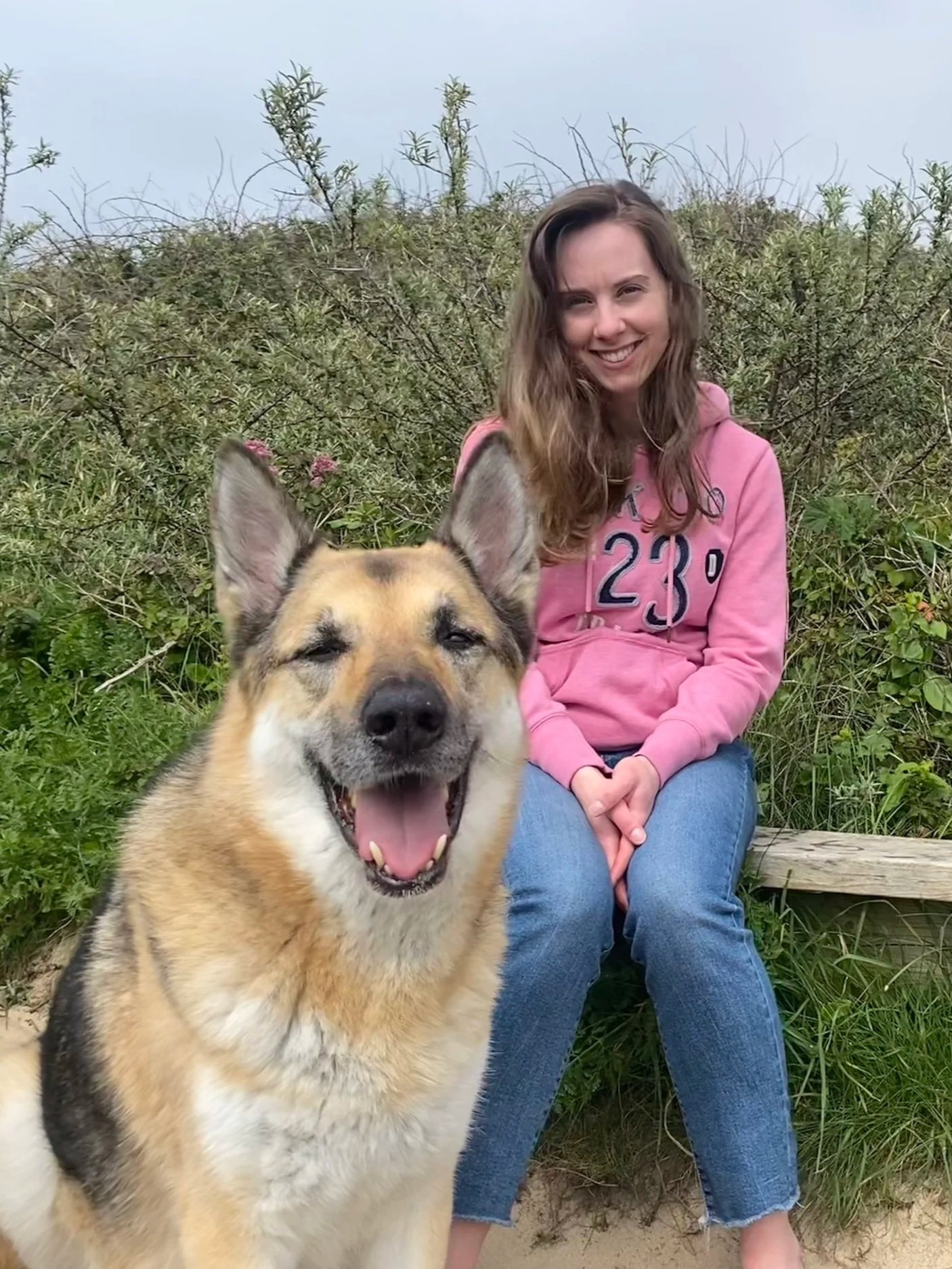 A young woman (Cheyenne) with long brown hair, wearing a pink hoodie and blue jeans, sitting outdoors with greenery in the background. A large, happy dog with tan and black fur and pointed ears is in the foreground, facing the camera.