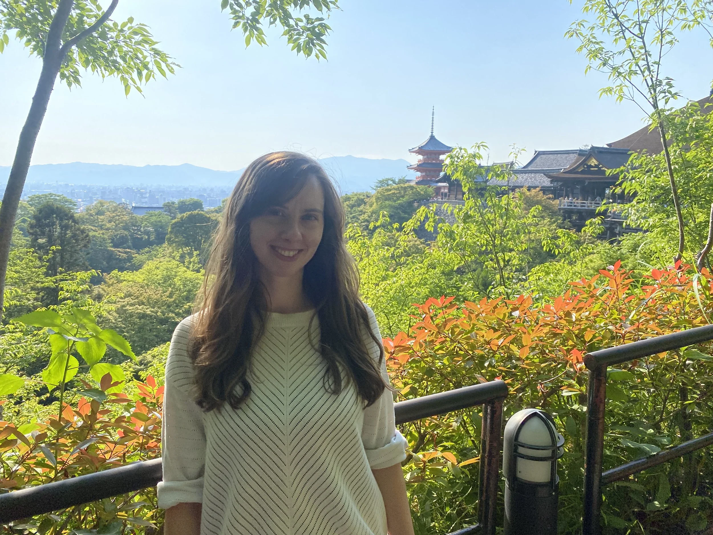A smiling woman with long dark hair standing outdoors among green foliage with traditional Japanese buildings and a pagoda in the background.