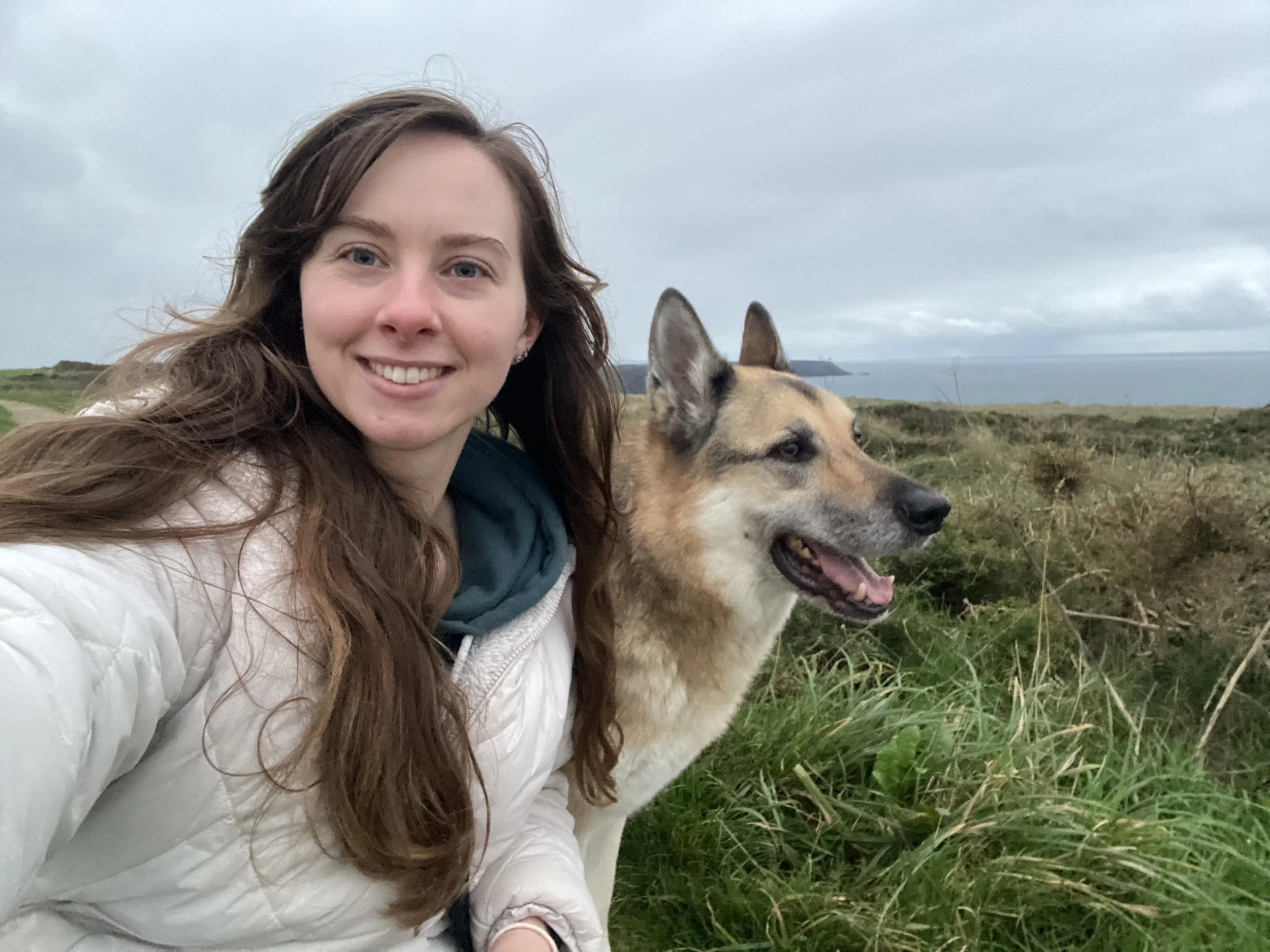 A smiling young woman with long brown hair taking a selfie outdoors on a cloudy day, with a medium-sized dog with upright ears, standing next to her in a grassy landscape overlooking the ocean.