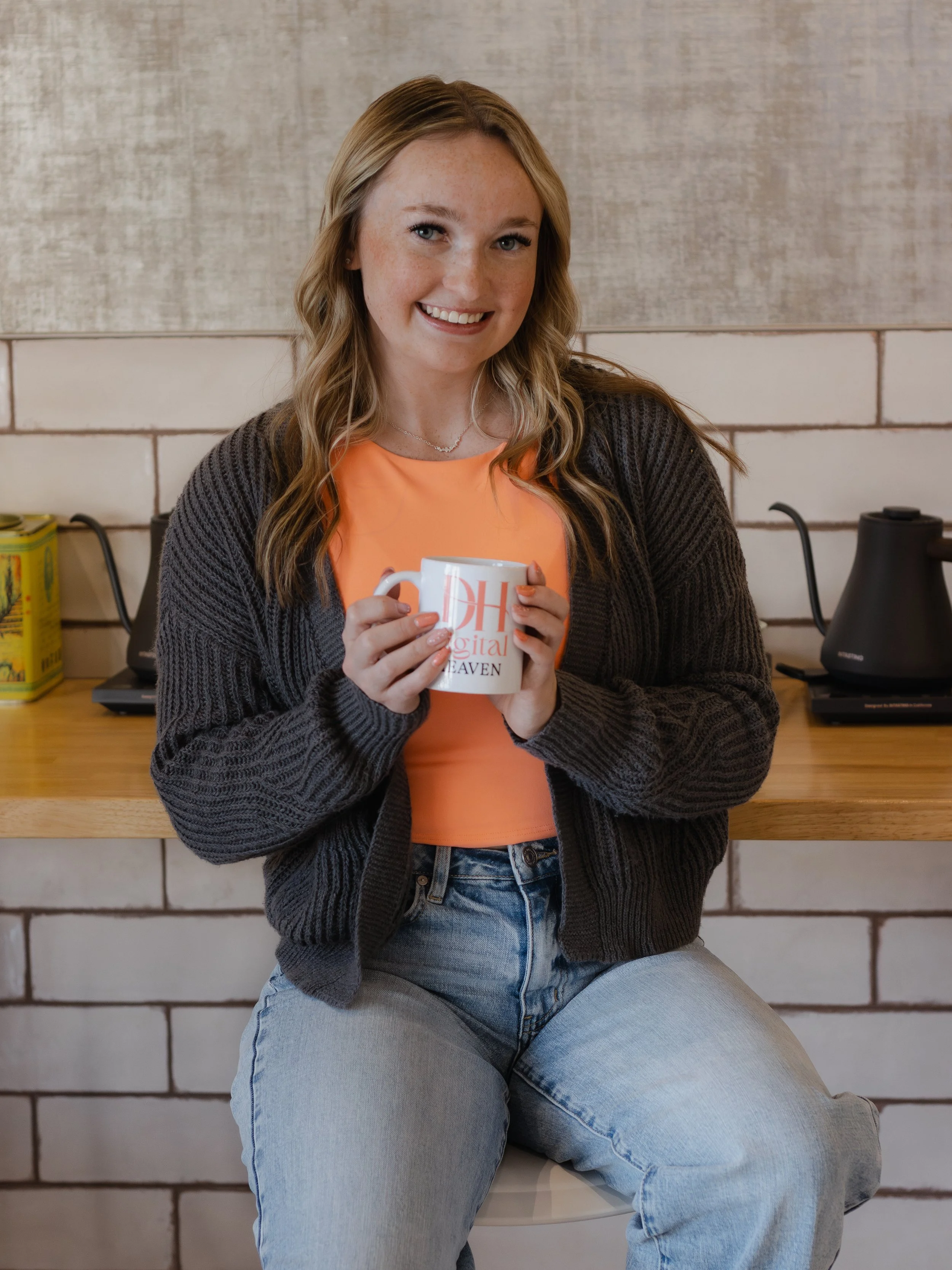 Nevaeh owner of Digital Heaven, wearing a black cardigan, coral top, and light blue jeans, sitting on a stool in front of a brick wall. She is smiling and holding a mug with custom Digital Heaven logo on it.