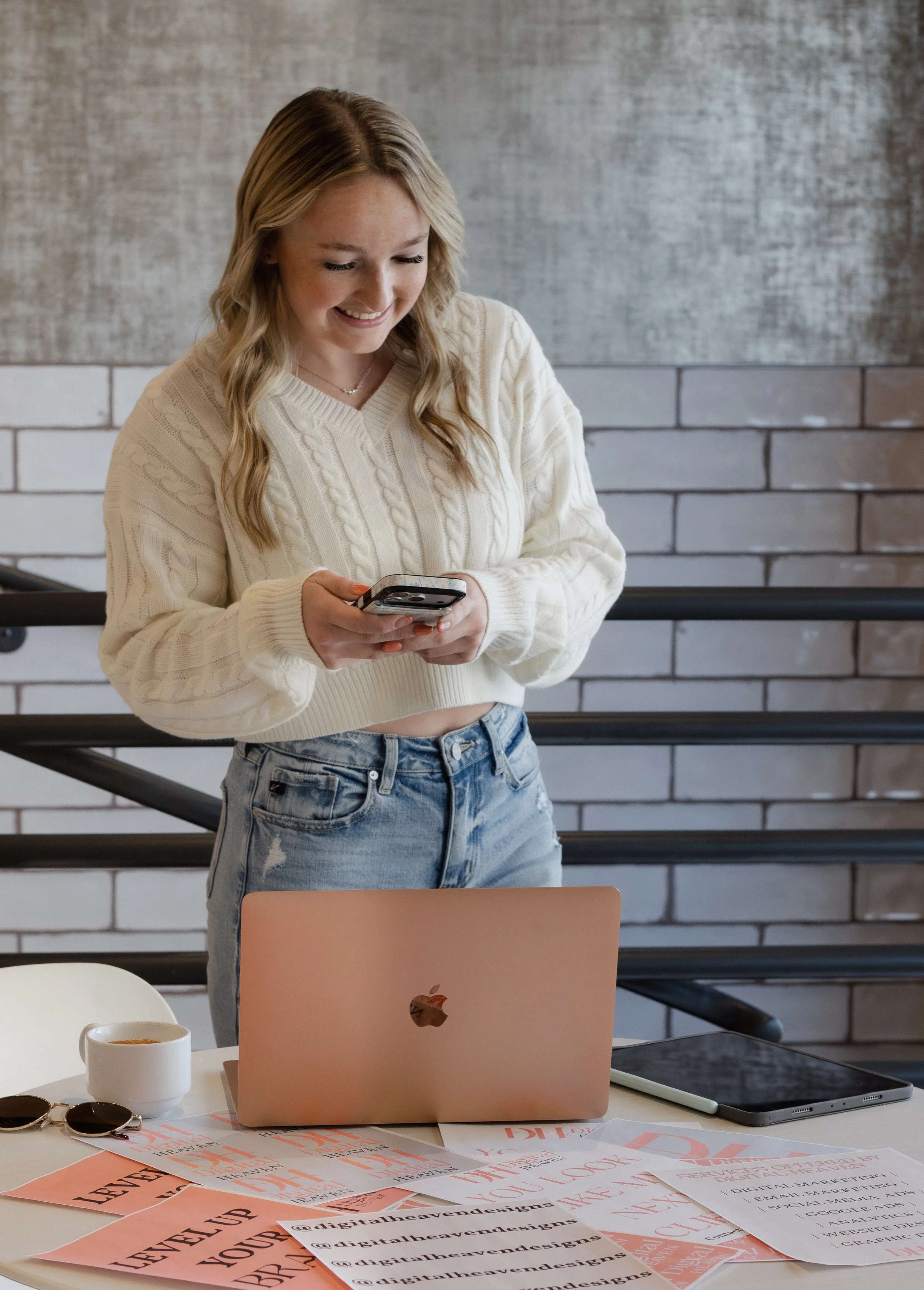 Nevaeh owner of Digital Heaven wearing a cream-colored sweater and jeans is smiling and looking at her phone. On the table in front of her, there are various papers, a laptop, a tablet, sunglasses, and a white Digital Heaven mug.