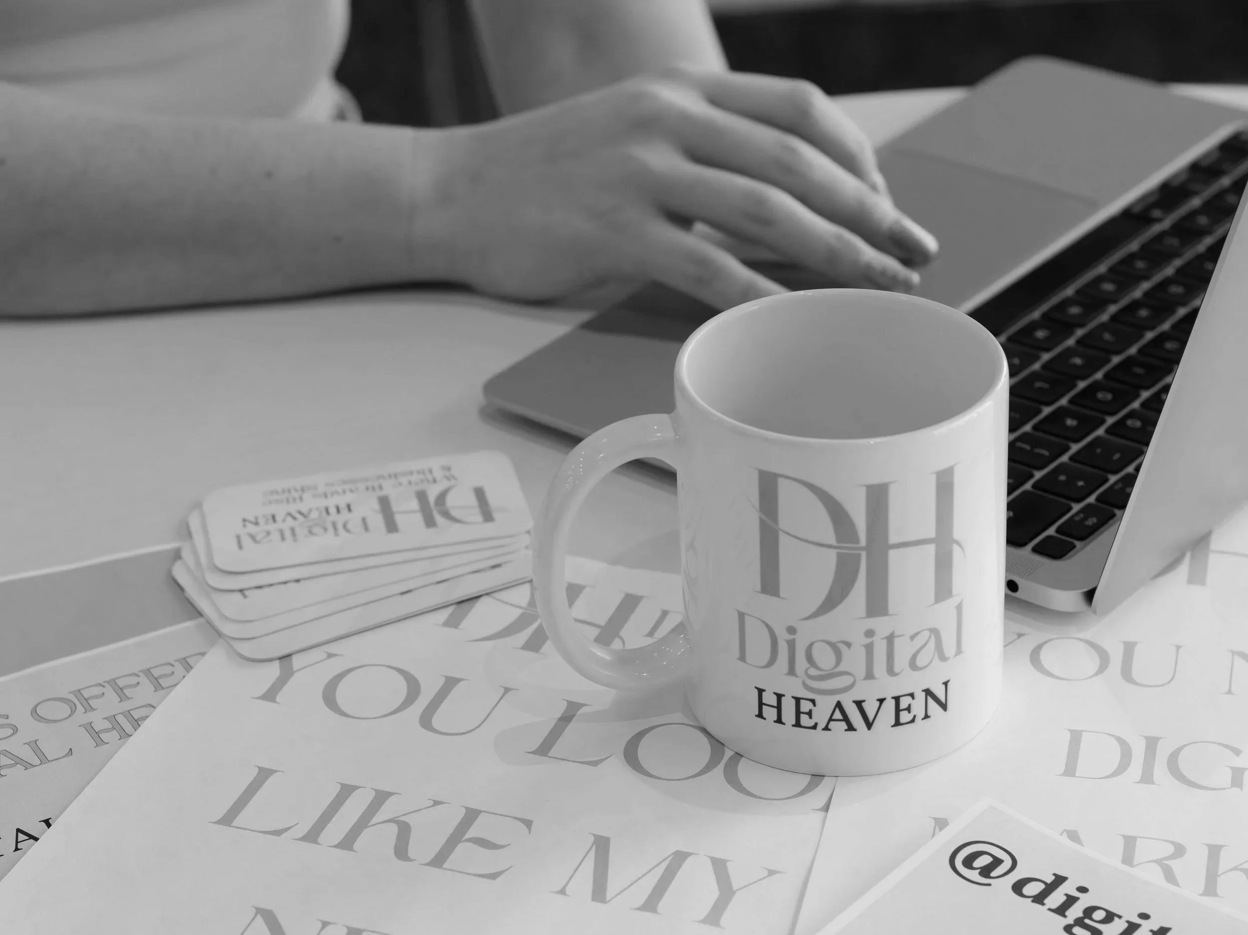 Nevaeh owner of Digital Heaven typing on a laptop with a custom white Digital Heaven coffee mug on the table. Several paper cards with printed words are scattered on the table, partially visible.