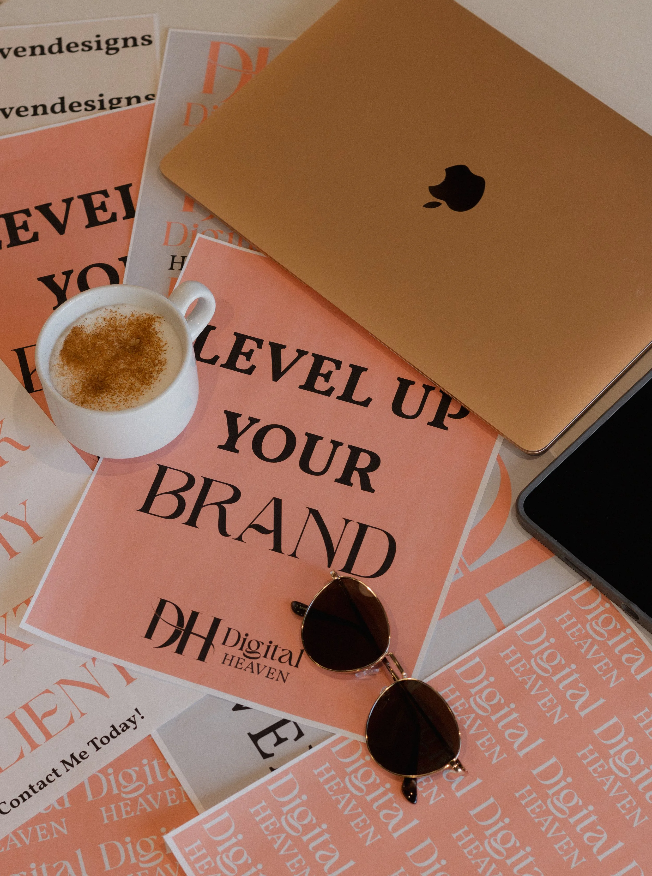 A collection of pink and white flyers and posters with the words 'LEVEL UP YOUR BRAND' and 'Digital HEAVEN' printed on them, a gold laptop with a black Apple logo, a cup of coffee with cinnamon on top, and sunglasses placed on the flyers.