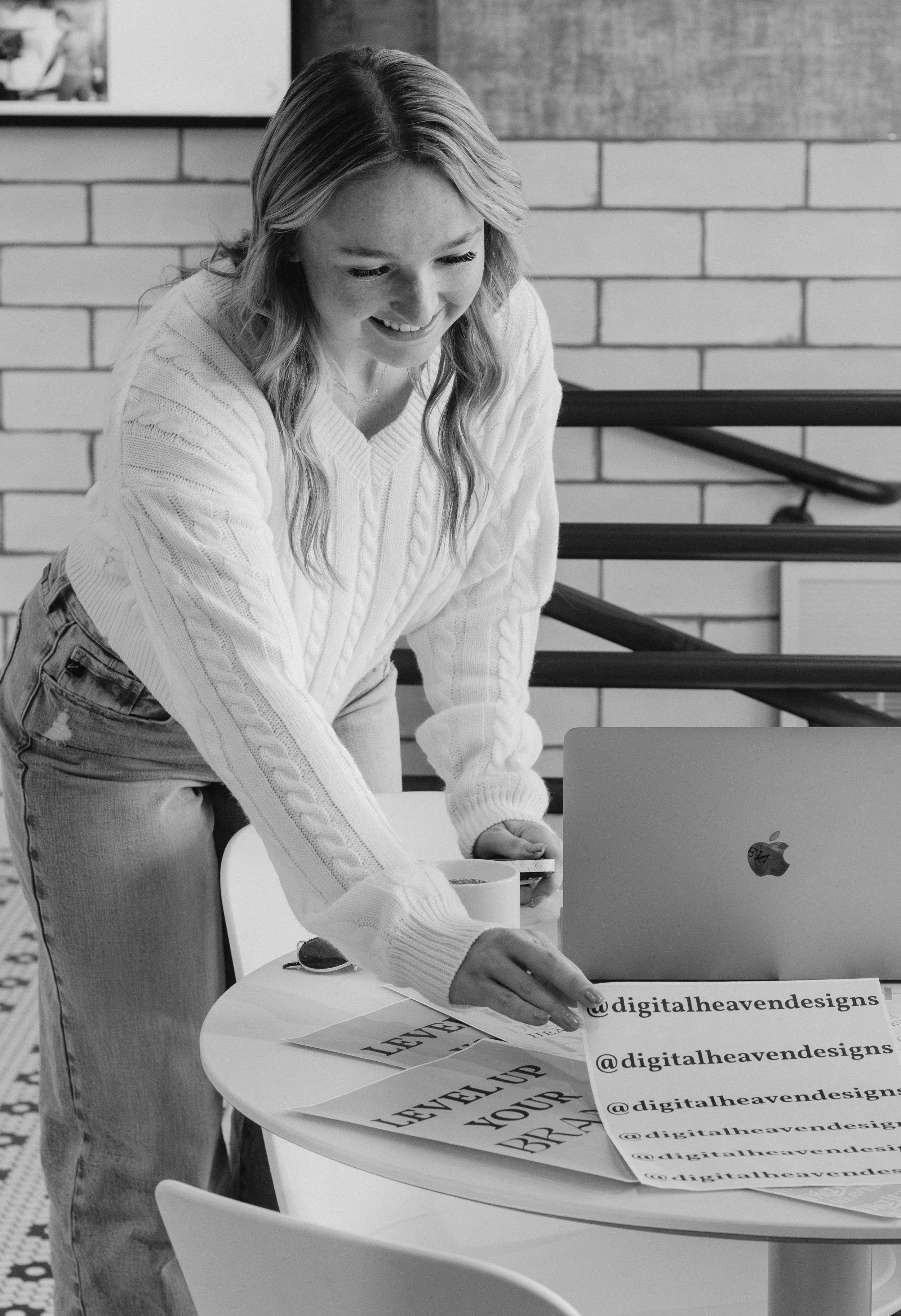Nevaeh owner of Digital Heaven wearing a sweater and jeans standing at a round table, looking at printed text and a laptop with social media handles on posters, in a modern office with brick walls.