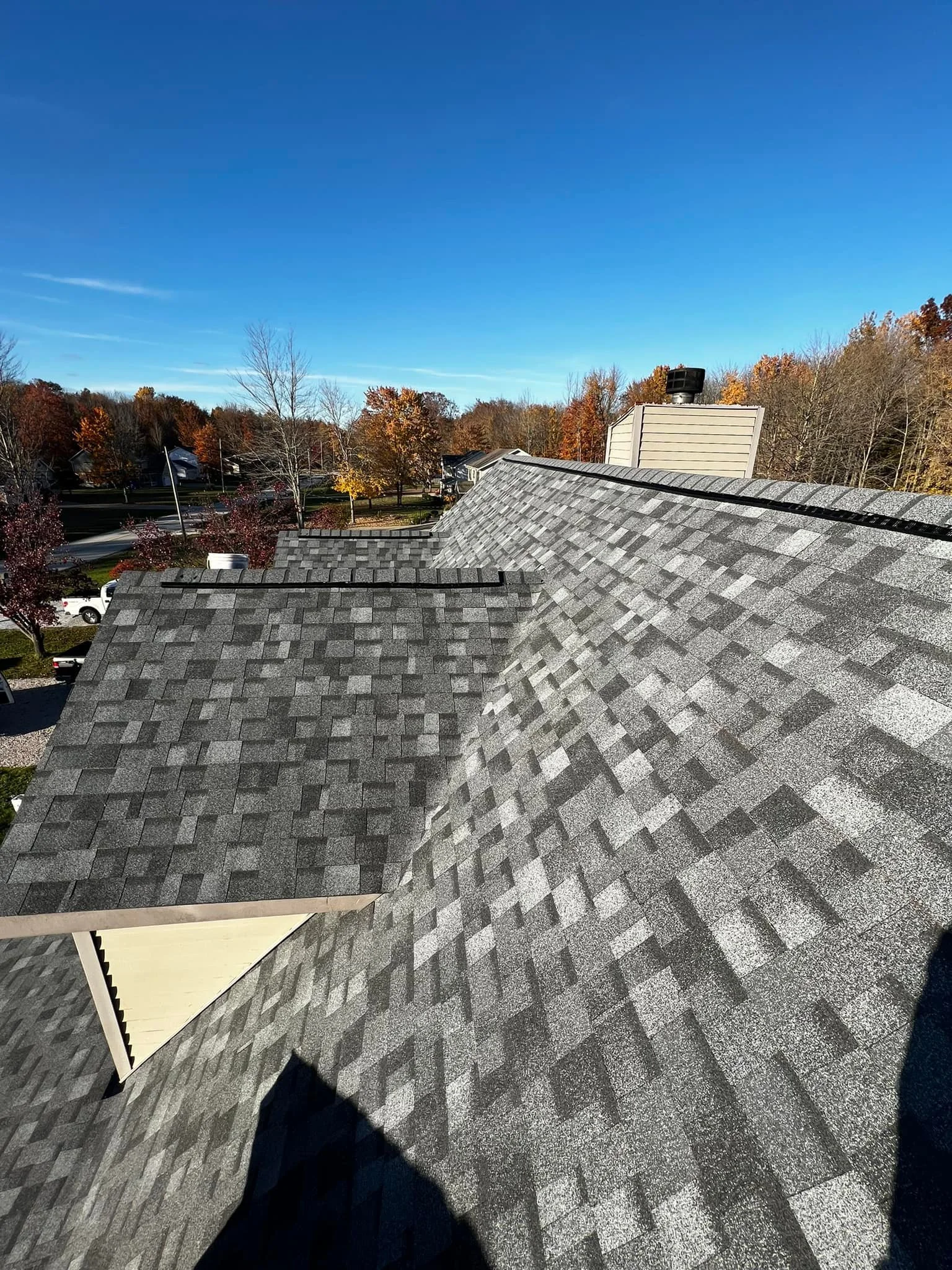 View from a rooftop showing asphalt shingles, a vent, and trees with fall foliage in the background under a clear blue sky.