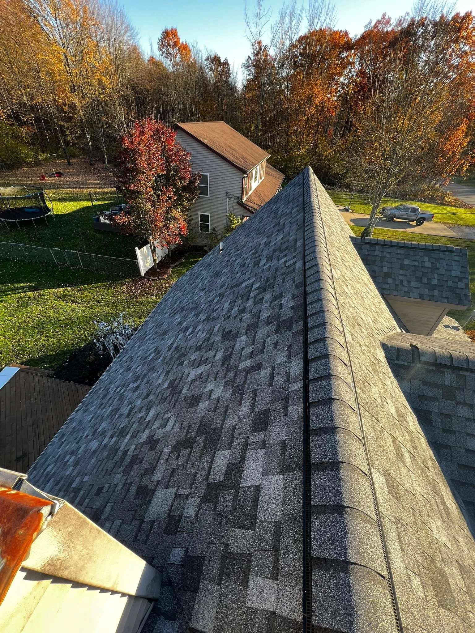 Aerial view of a roof with asphalt shingles on a house, surrounded by trees with autumn foliage, a yard with grass, and a parked truck.