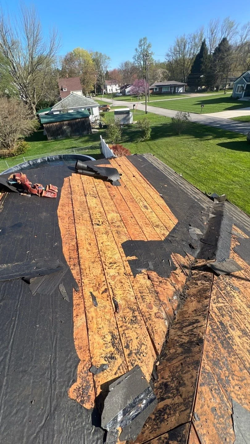 View of a house roof with damaged black underlayment and missing shingles, revealing the wood underneath, in a suburban neighborhood during daytime.