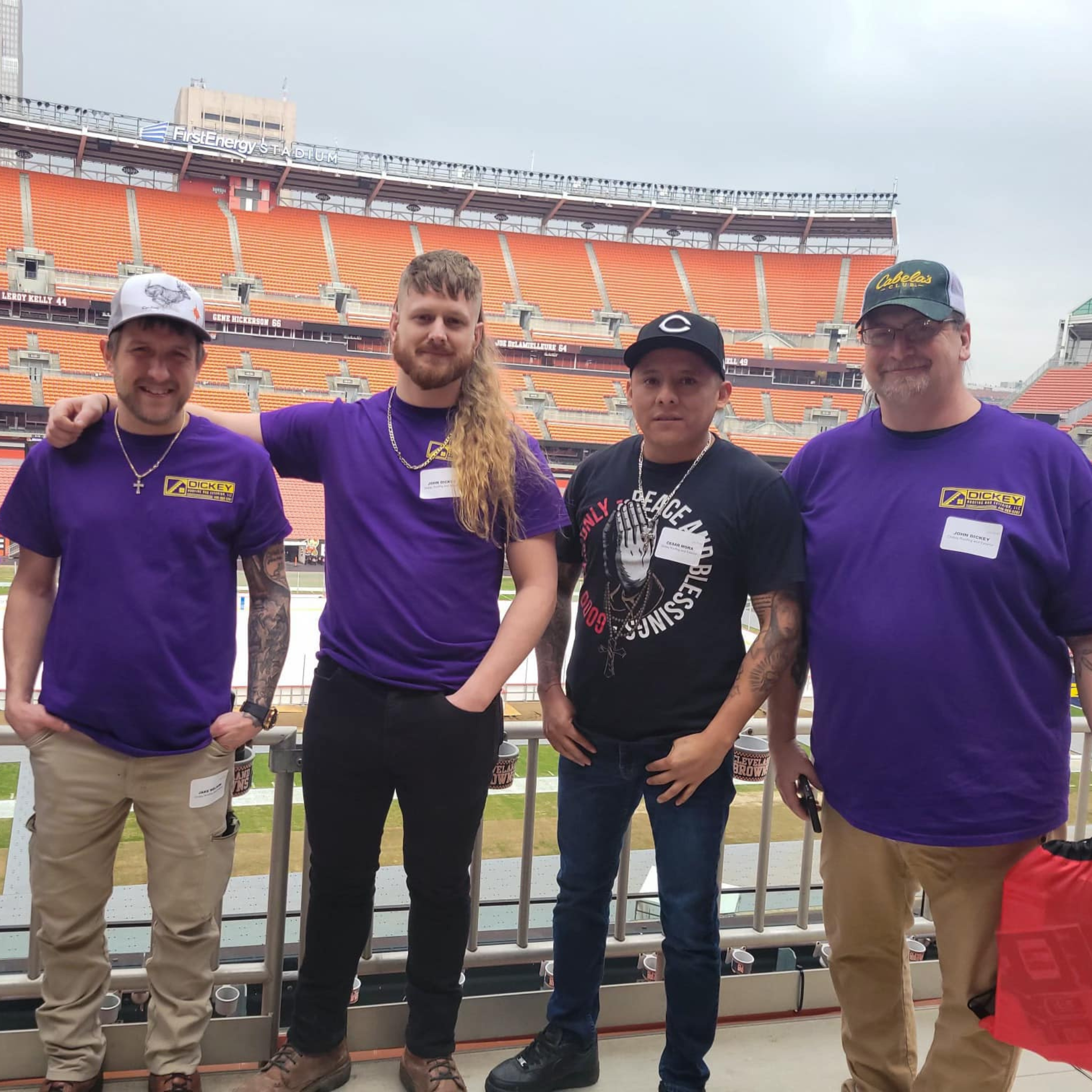 Four men standing together in a stadium, two wearing purple shirts with a logo, one wearing a black T-shirt, and one wearing a white cap, with an empty stadium in the background.