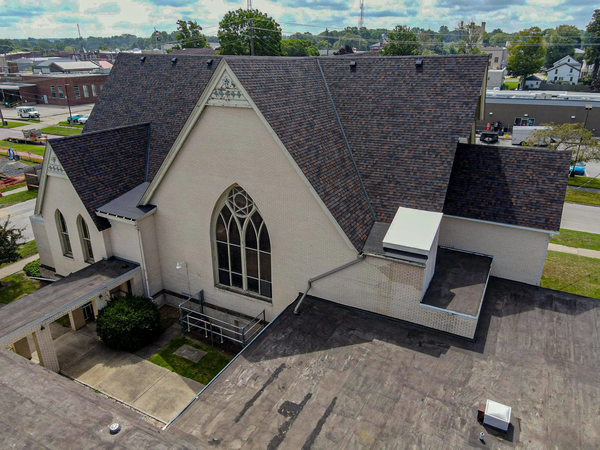 Aerial view of a church with a steep gable roof, large stained glass window, and white brick walls. Surrounding buildings and streets are visible in the background.
