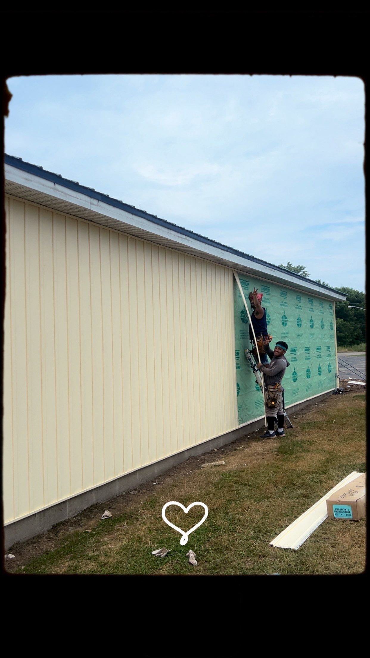 Two construction workers installing siding on the exterior of a building, one on a ladder waving, the other holding the ladder.