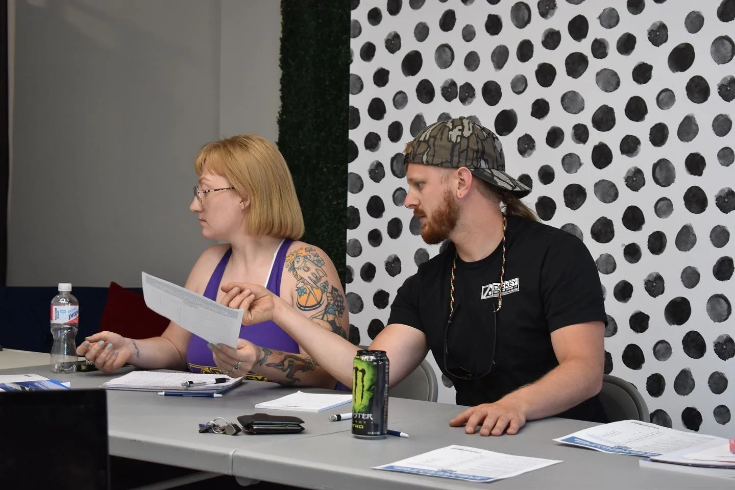 Two people sitting at a table in a meeting room, one woman wearing glasses and a tank top with tattoos on her arm, and a man with a beard wearing a camouflage cap and a black T-shirt, handing a document to the woman. The table has notebooks, a water bottle, a can of Monster energy drink, a phone, and pens.