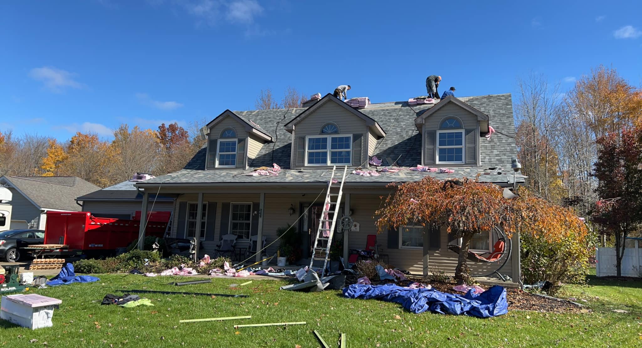 Two workers on the roof of a house under construction, with materials and tools scattered on the roof and yard, in a suburban neighborhood with trees showing fall foliage.
