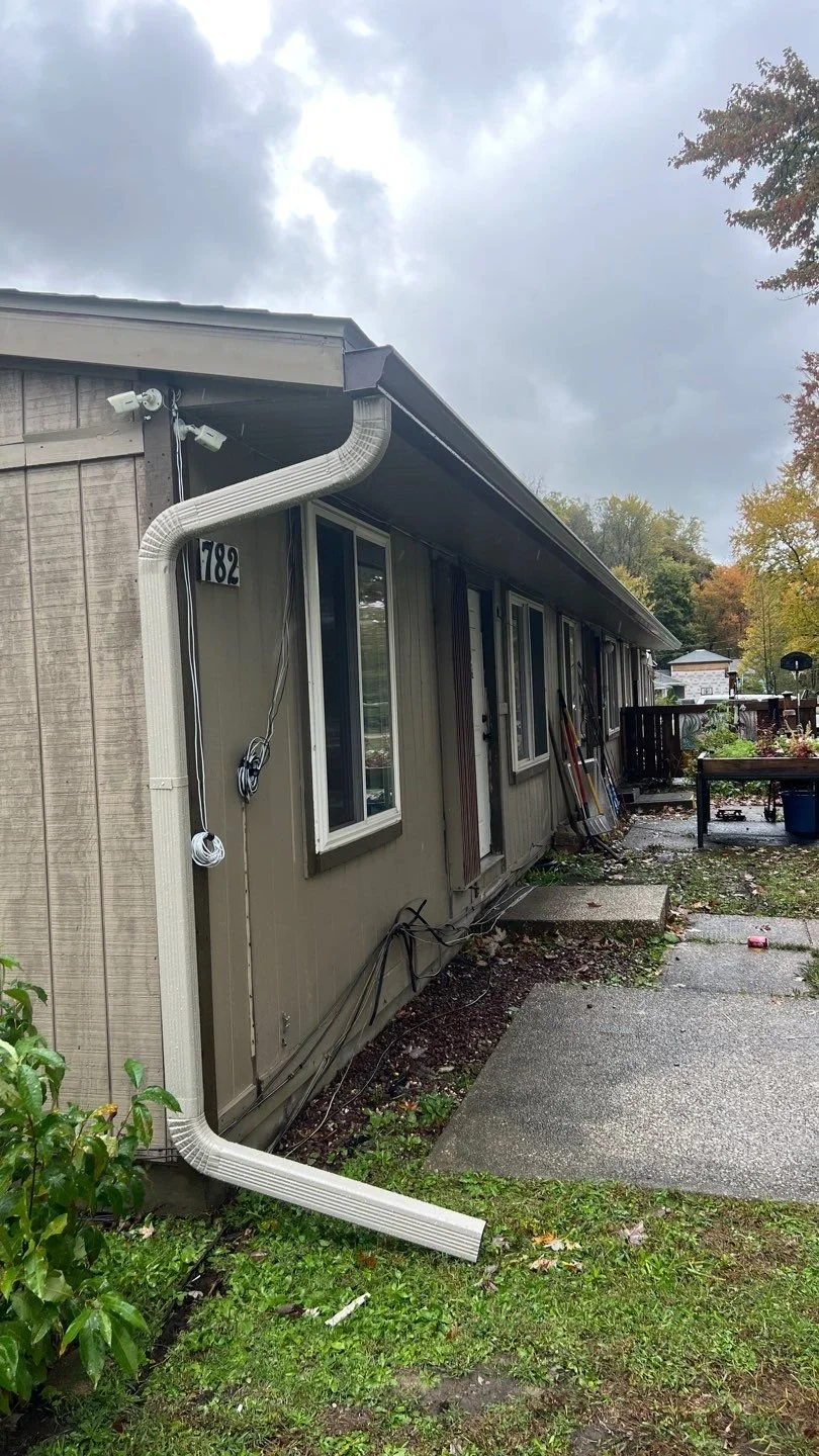 Side view of a house with a downspout pipe disconnected and hanging near the ground, on a cloudy day.