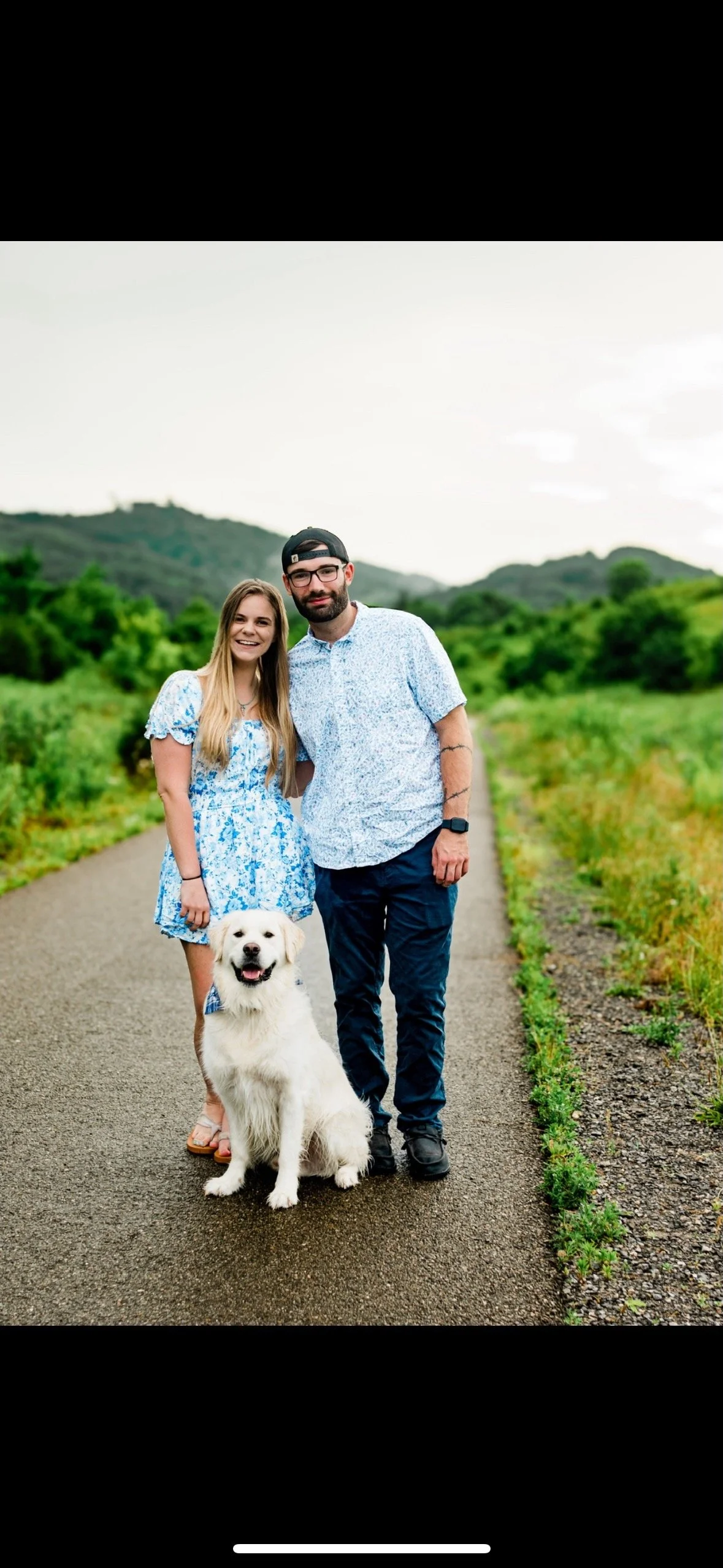 A smiling woman in a blue floral dress, a bearded man in a light blue shirt and dark pants, and a large white dog, all standing on a paved path in a green outdoor area with hills in the background during daytime.