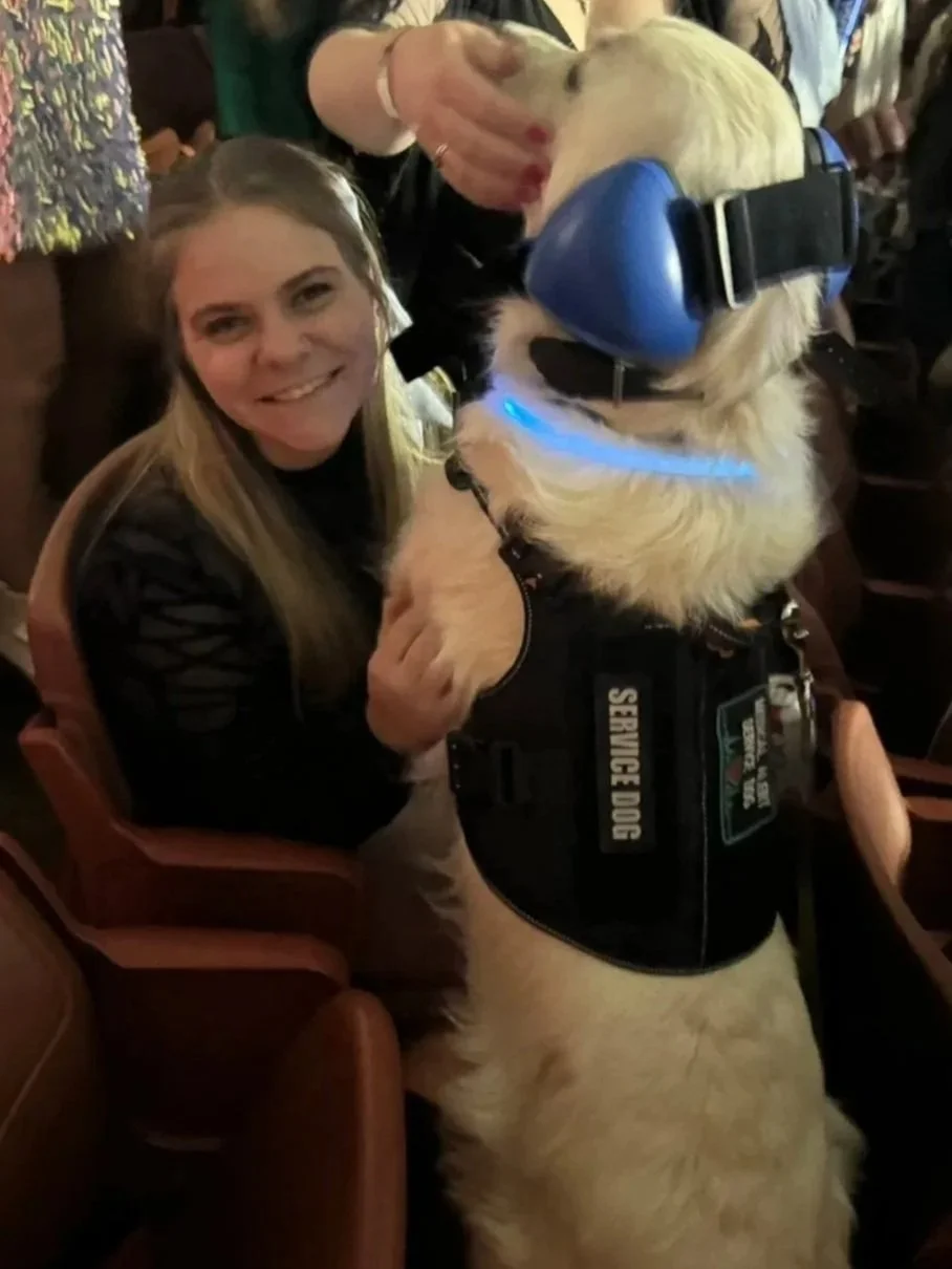 A woman smiling while holding a service dog wearing noise-canceling headphones and a vest that says 'SERVICE DOG'. The dog is a large, light-colored retriever, and they are seated in a crowded indoor setting.